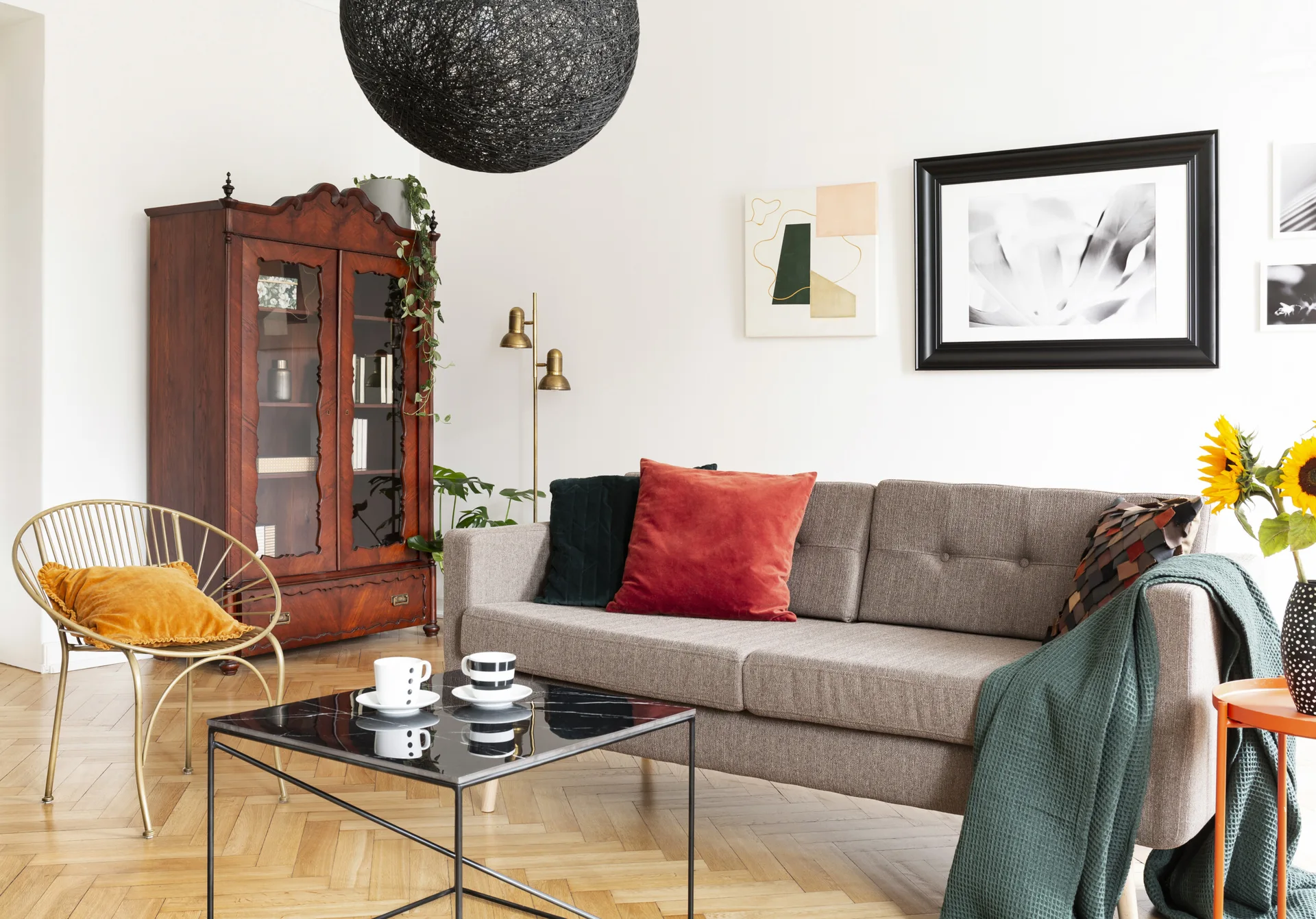 An eclectic living room features a grey sofa, a black marble coffee table, a tall vintage wooden cabinet, and herringbone parquet wood flooring.
