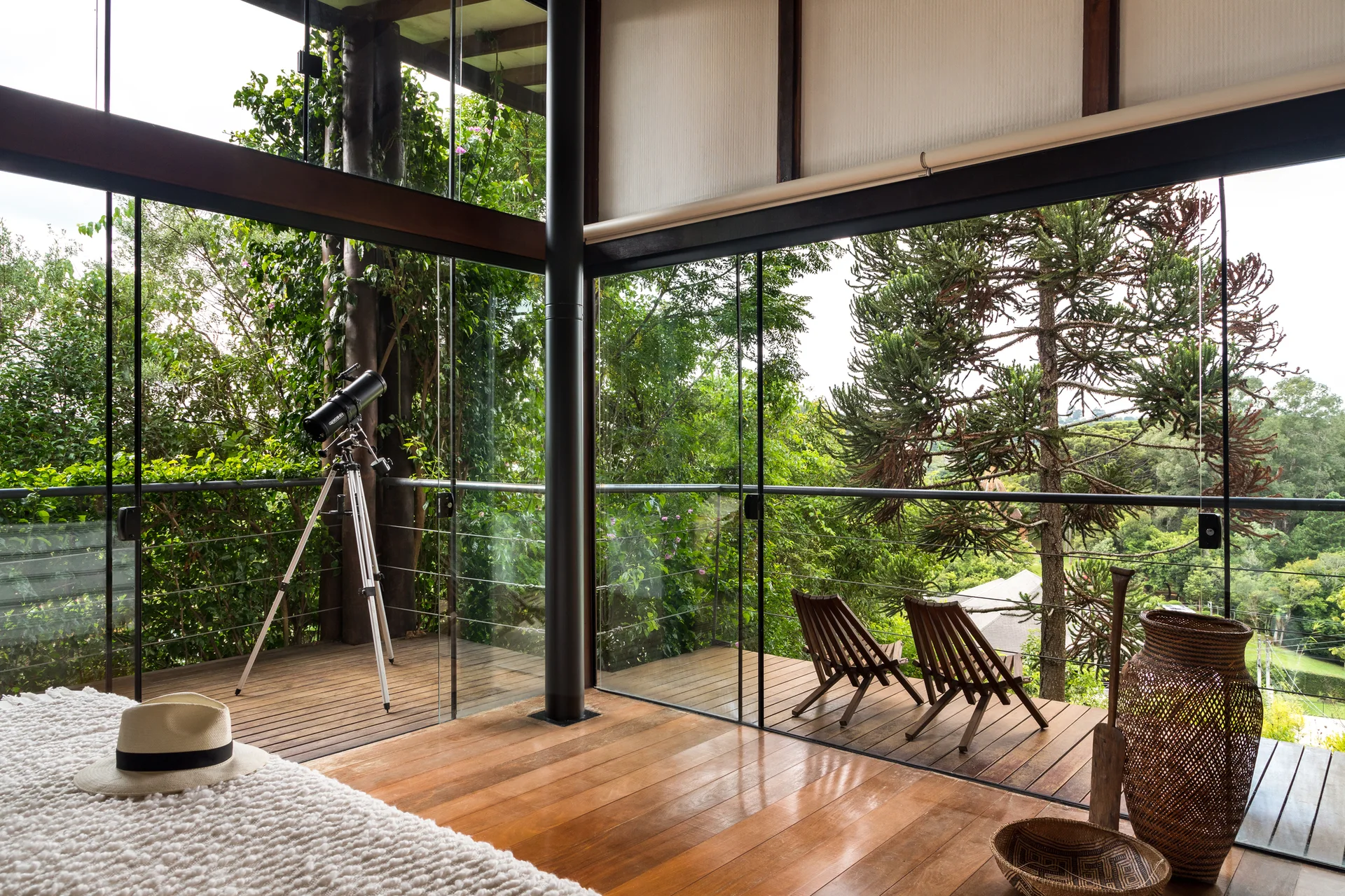 A bedroom with wood floors and glass walls looks out onto an outdoor wooden deck with lounge chairs and a telescope surrounded by trees.