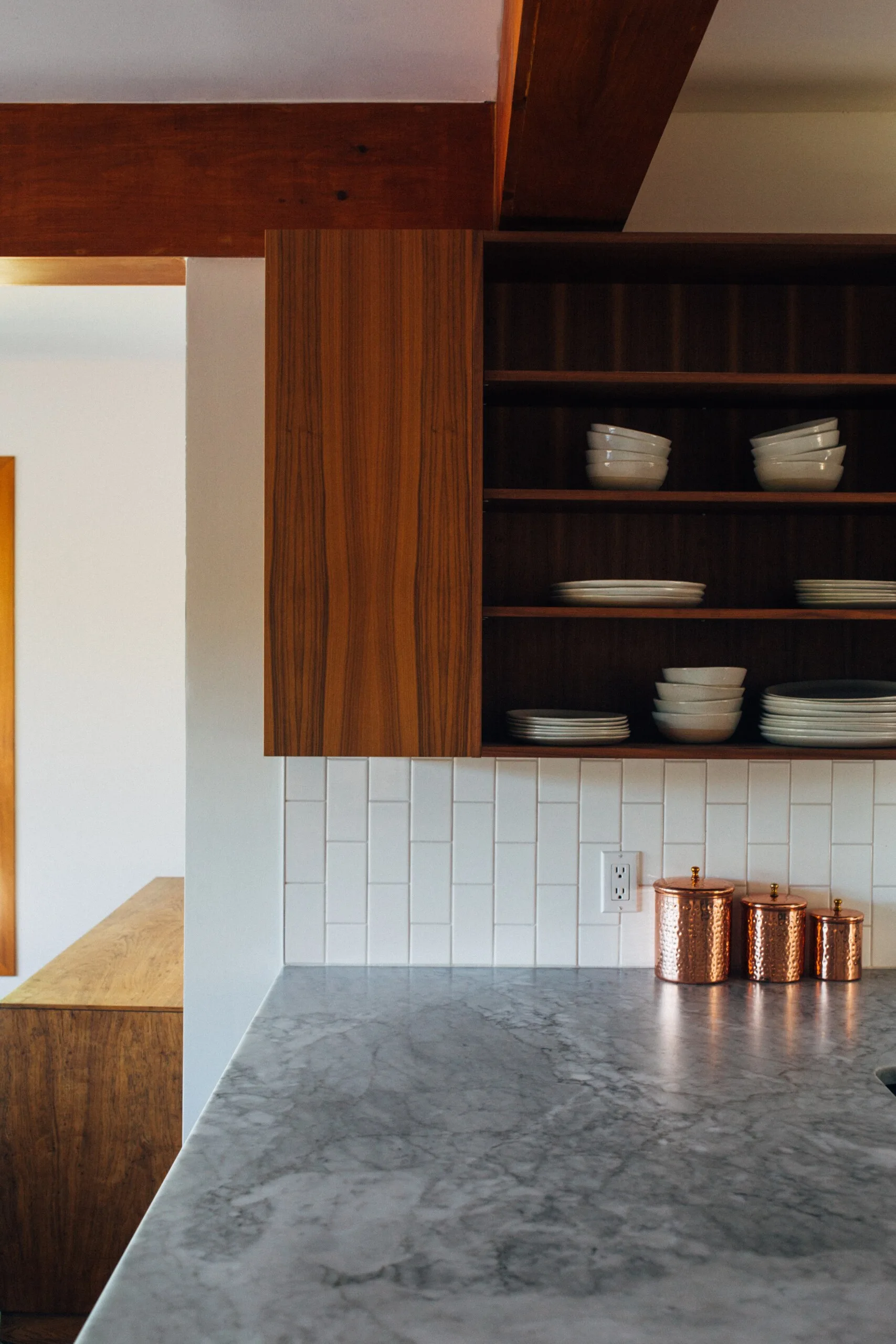 A kitchen corner featuring dark wood cabinets with open shelving, a vertical white subway tile backsplash, a grey marble countertop, and copper canisters.
