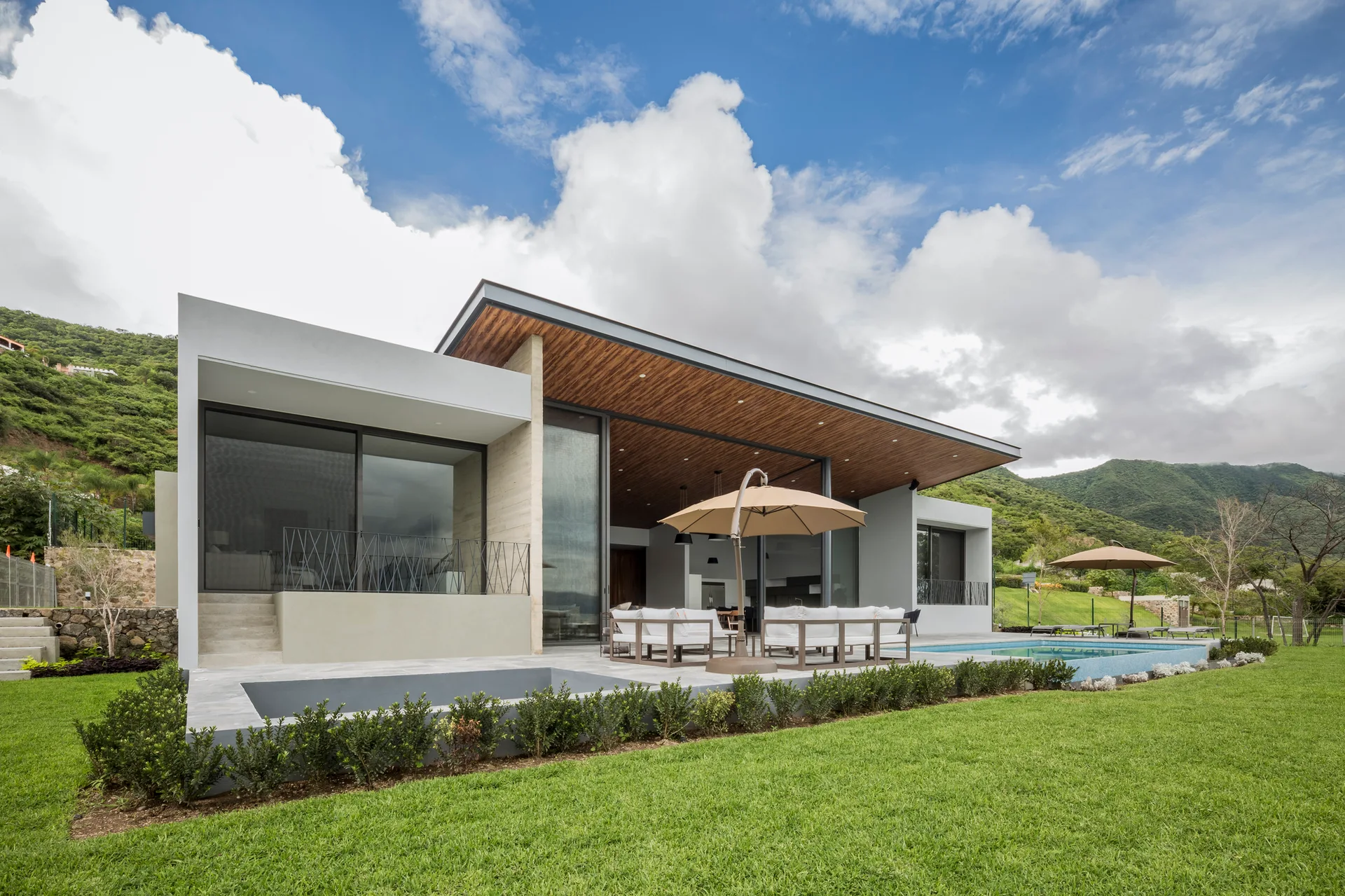 A modern concrete home features a gray stone patio with white outdoor furniture, a tan umbrella, a swimming pool, and a slanted wood ceiling overhang.