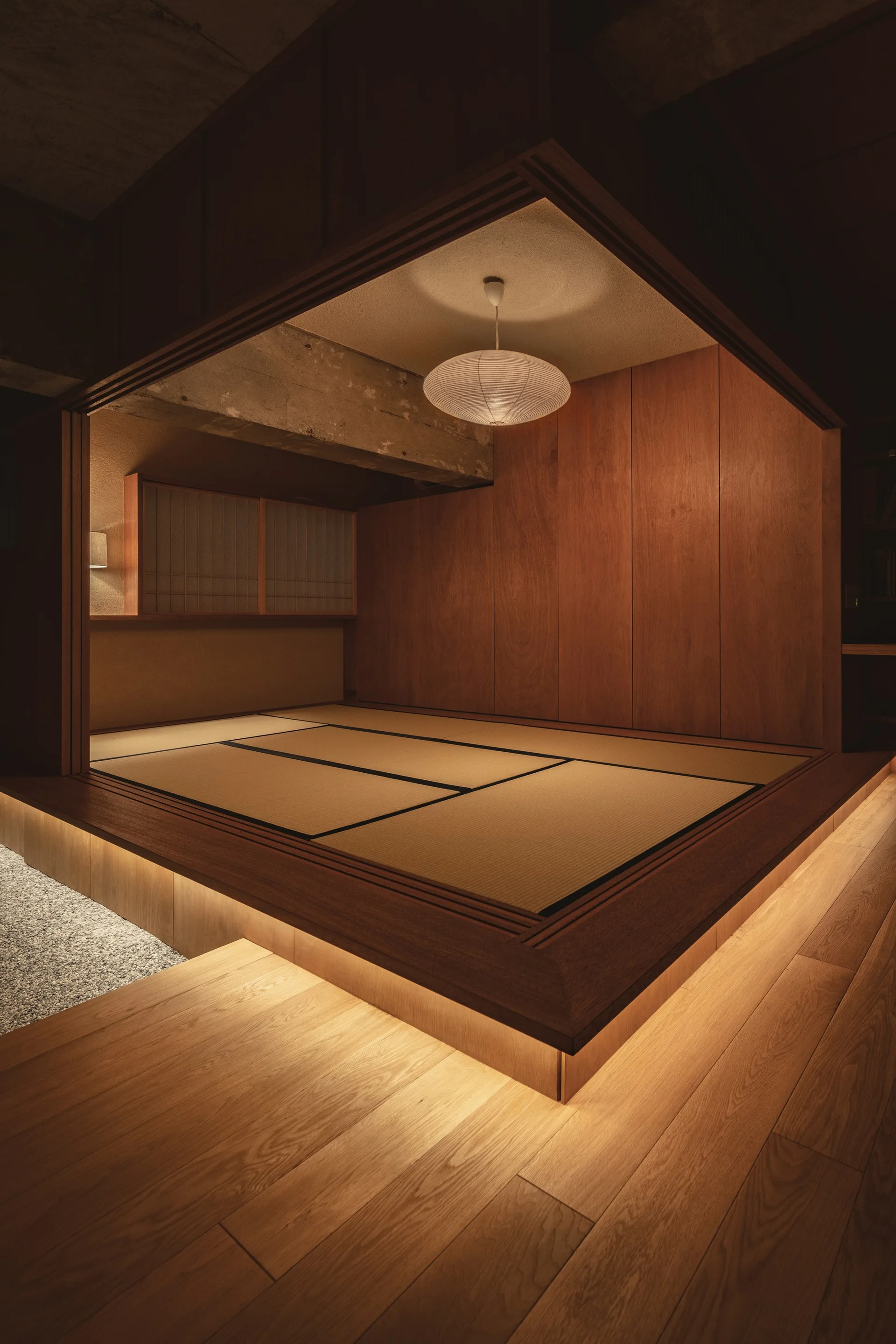 A raised wooden platform with tatami mats features warm under-platform lighting, vertical wood paneling, and a paper pendant light.