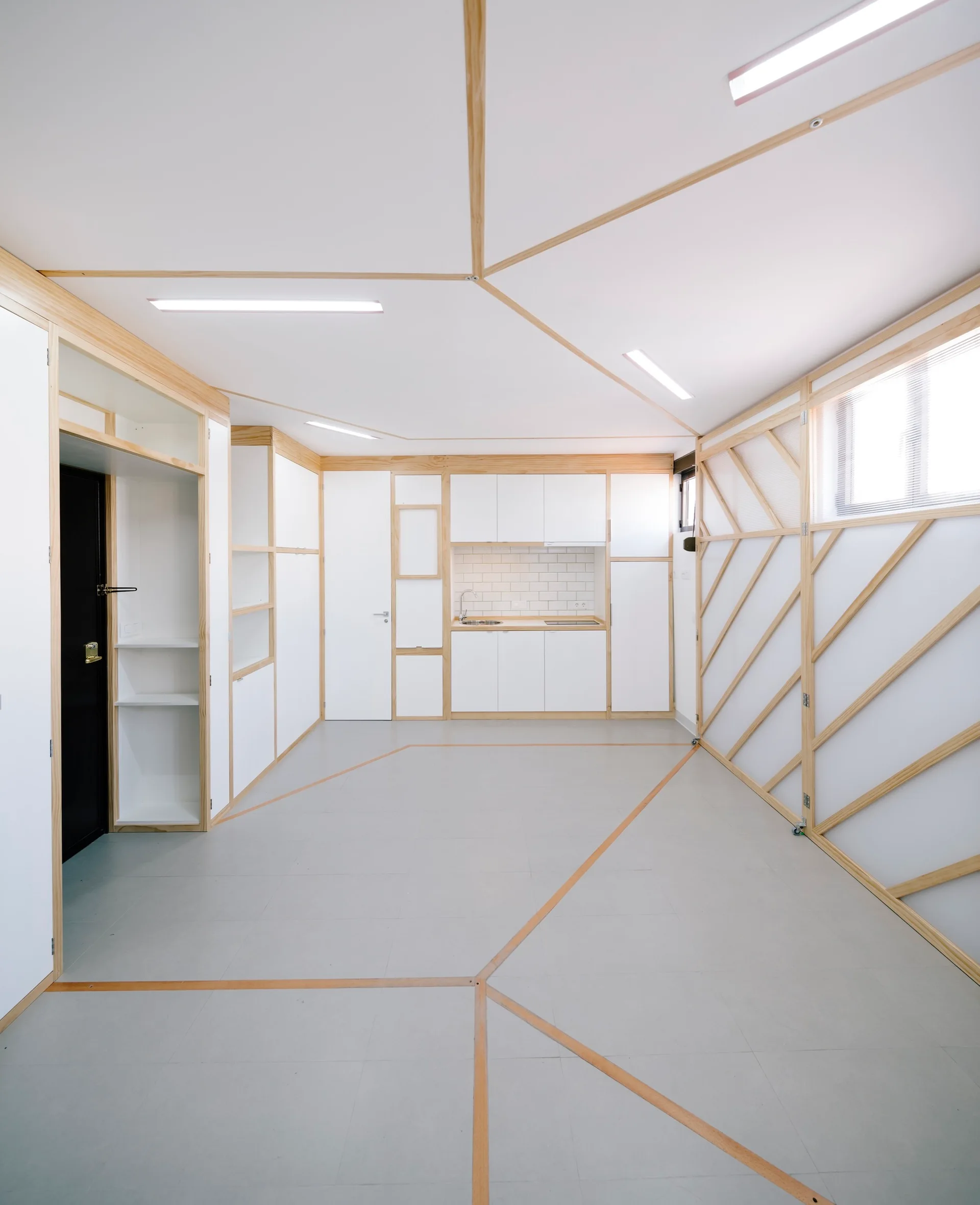A minimalist studio interior featuring white cabinets with light wood trim, linear ceiling lighting, and geometric wood lines on the floor and wall.
