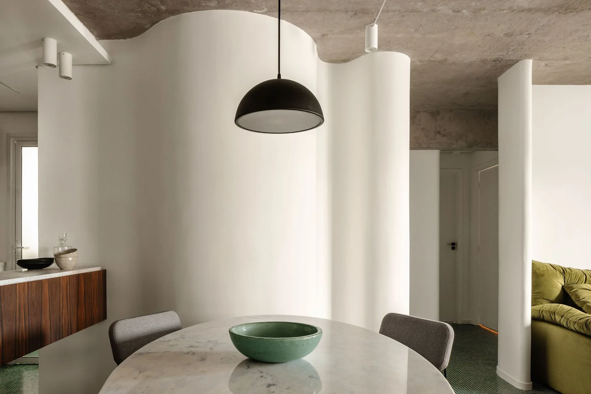 A minimalist dining area featuring an oval marble table, grey chairs, a black dome pendant light, and a prominent curved white feature wall.