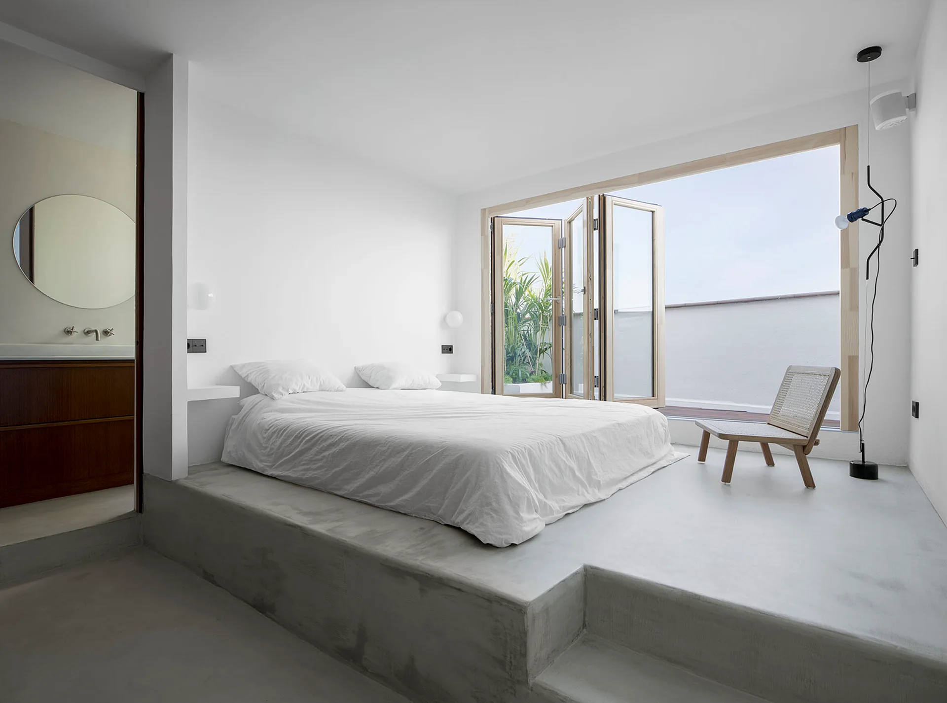 A minimalist bedroom featuring a bed on a raised grey concrete platform next to folding glass doors leading to a terrace.