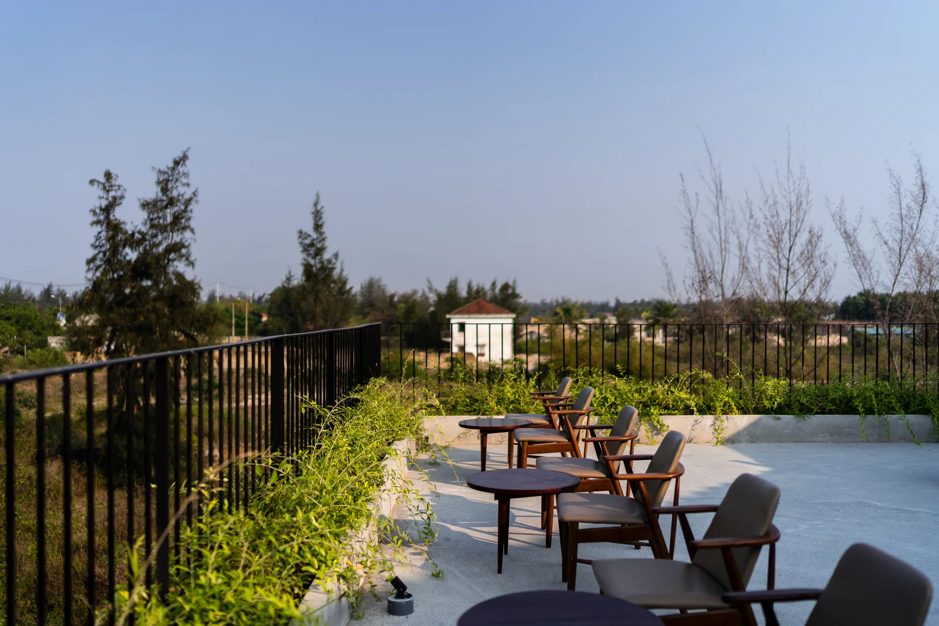 An open-air concrete terrace features several wood-framed armchairs and small round tables arranged next to a black metal railing and a low green planter.