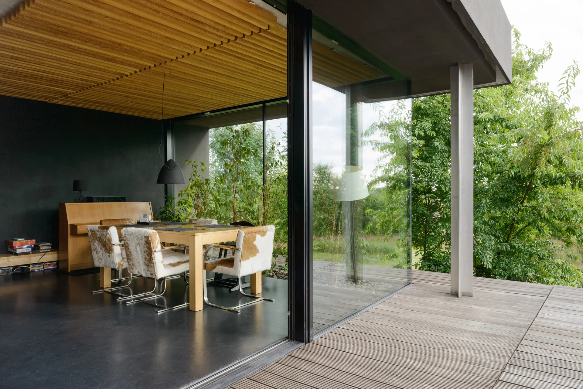A modern dining room with a light wood table, cowhide chairs, and a slatted wood ceiling, opening onto a wooden deck through sliding glass walls.
