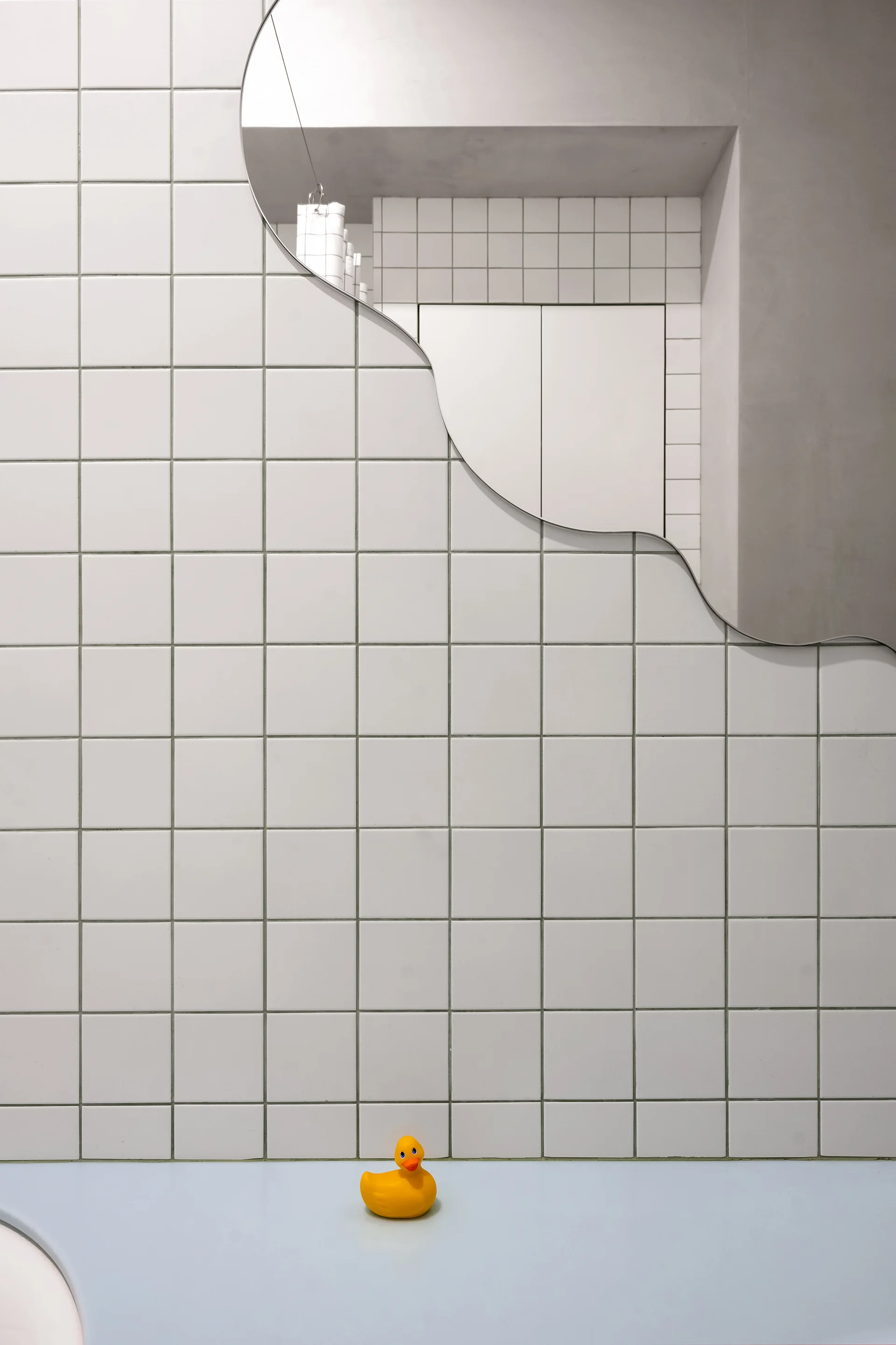 A minimalist bathroom wall featuring white square grid tiles, an organic curved mirror, and a yellow rubber duck sitting on a light blue ledge.