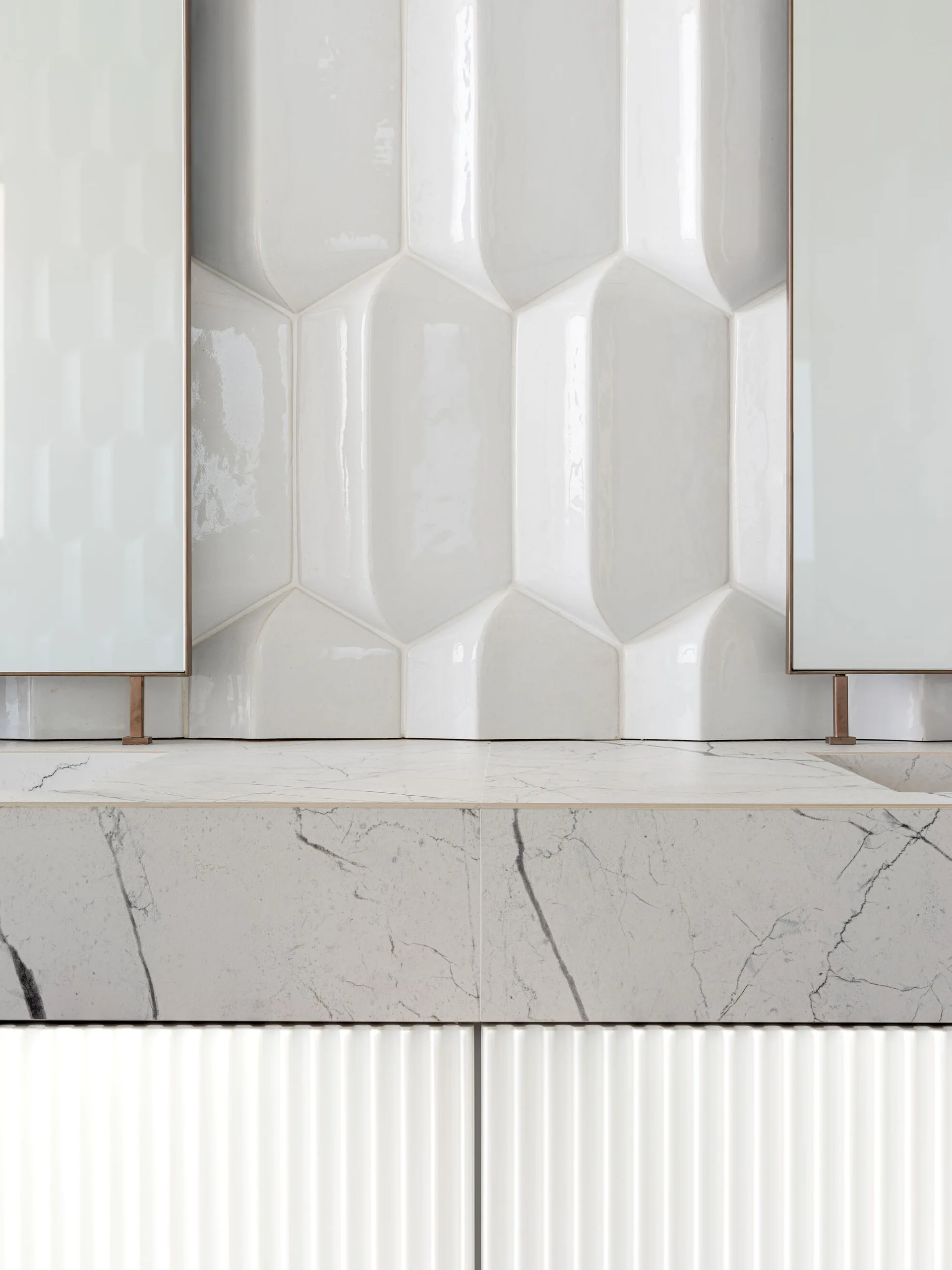 A close-up view of a bathroom vanity showing sculptural three-dimensional white wall tiles, a veined marble countertop, and fluted lower cabinetry.