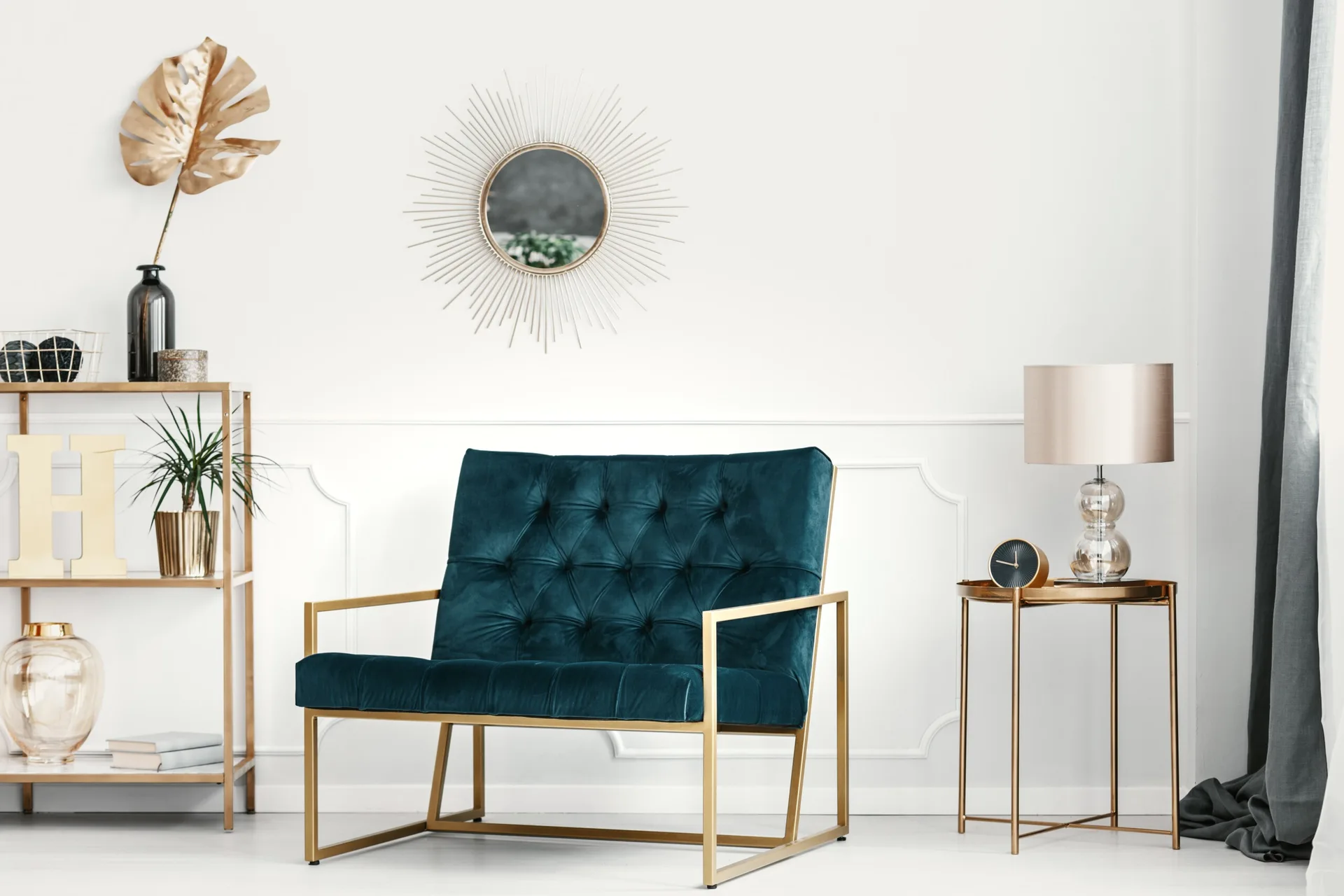 A teal velvet tufted armchair with a gold frame is flanked by a gold etagere and side table against a white wall with panel molding.