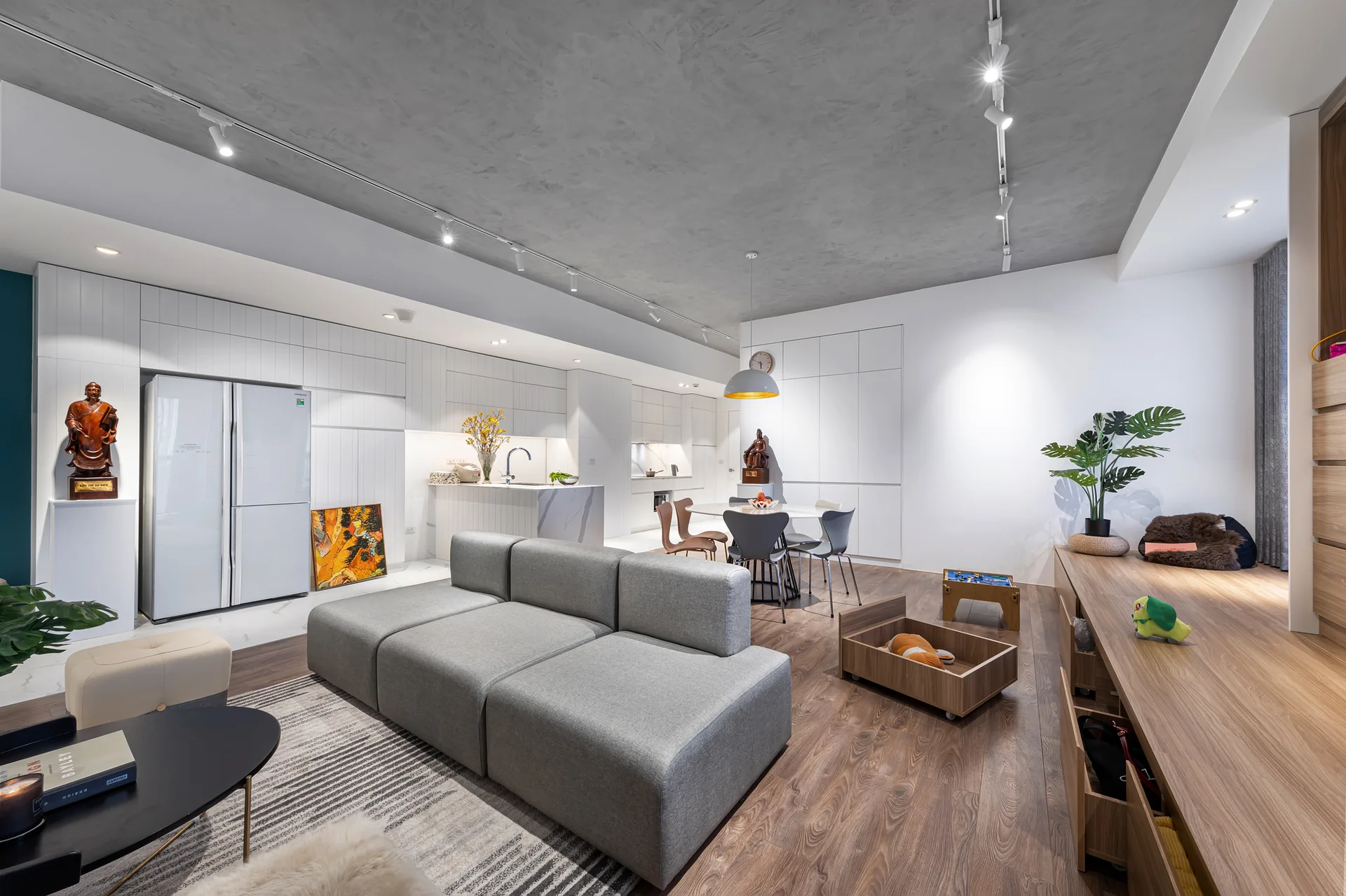 A modern open-plan living room and kitchen featuring a grey modular sofa, white flat-panel cabinetry, and a textured grey concrete ceiling.