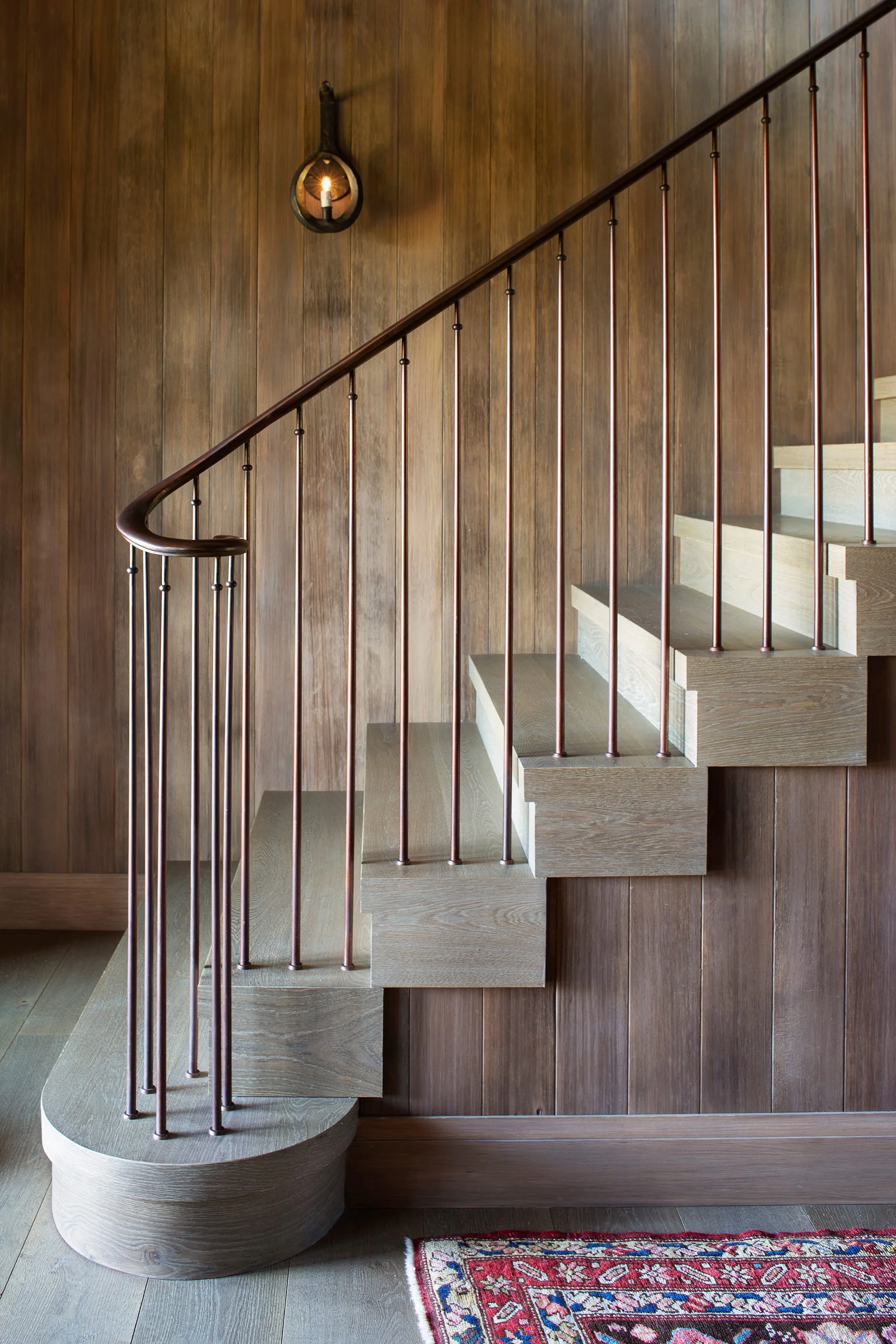 A wooden staircase features vertical wood paneling, dark metal spindles, and a curved bottom step beside a patterned area rug.