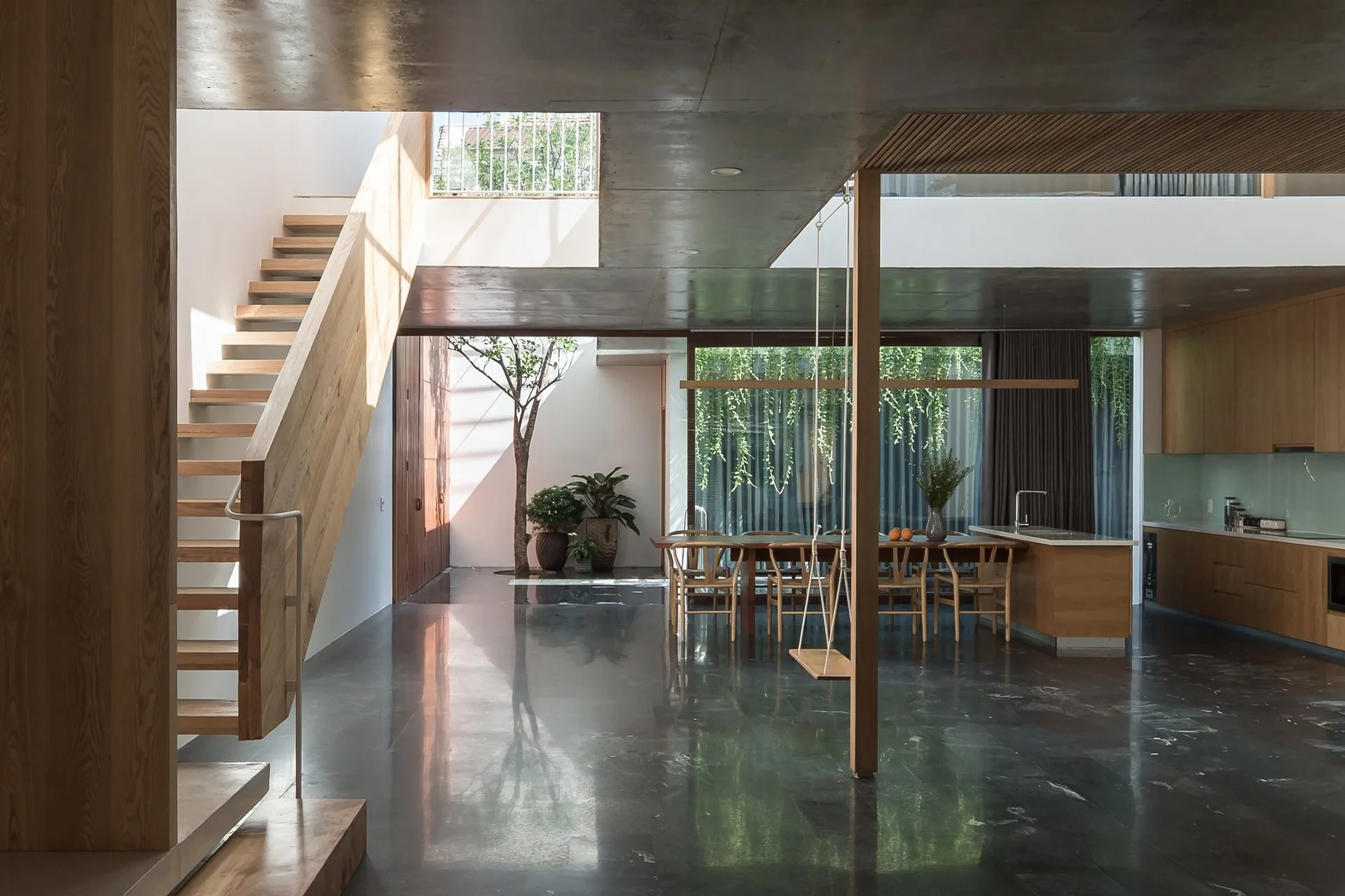 A spacious open-plan interior features a polished dark stone floor, a natural wood staircase, an indoor tree in an atrium, and a dining table.