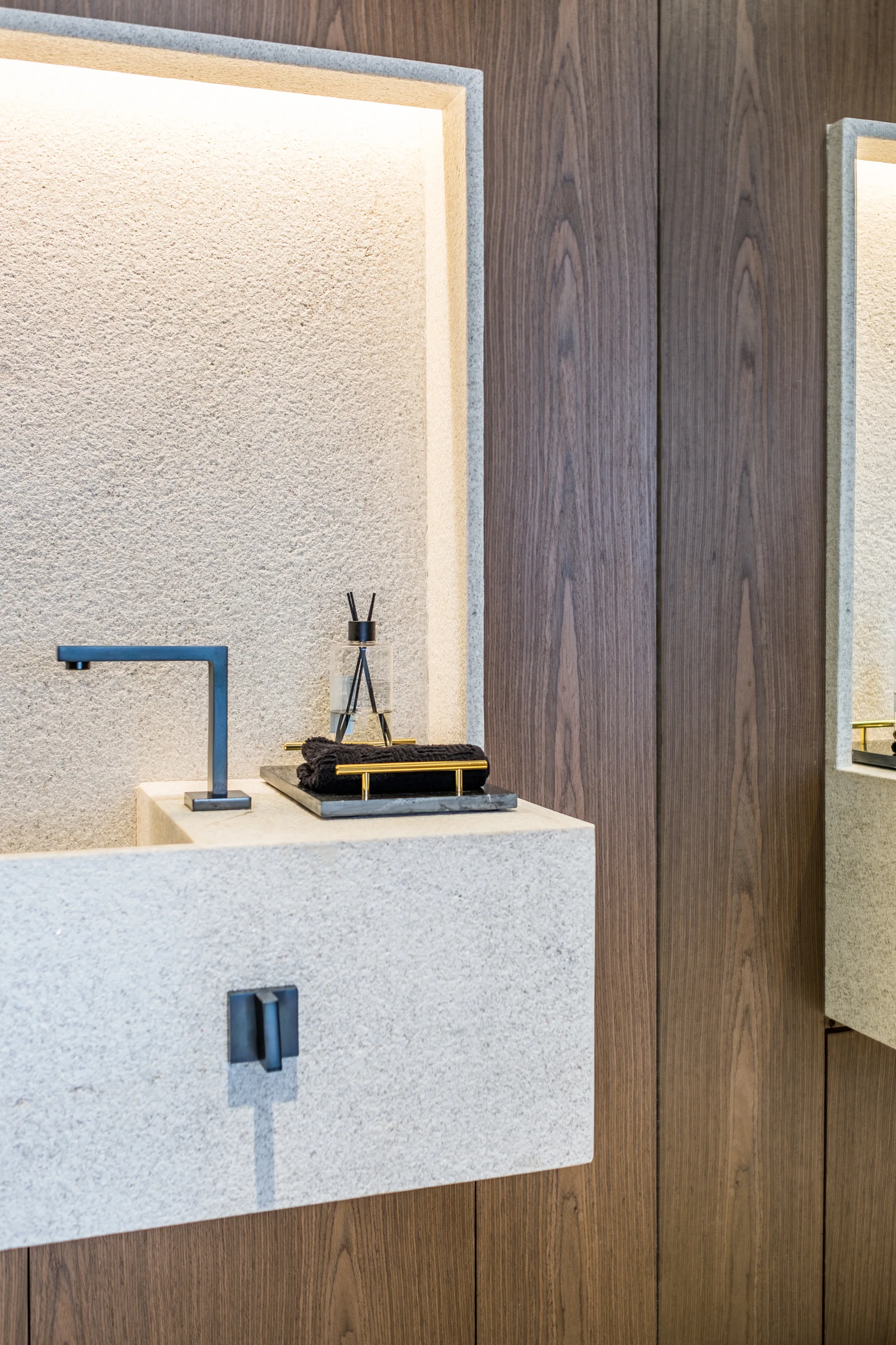 A minimalist textured stone washbasin features a matte black faucet set against a dark wood wall and a lit recessed niche.