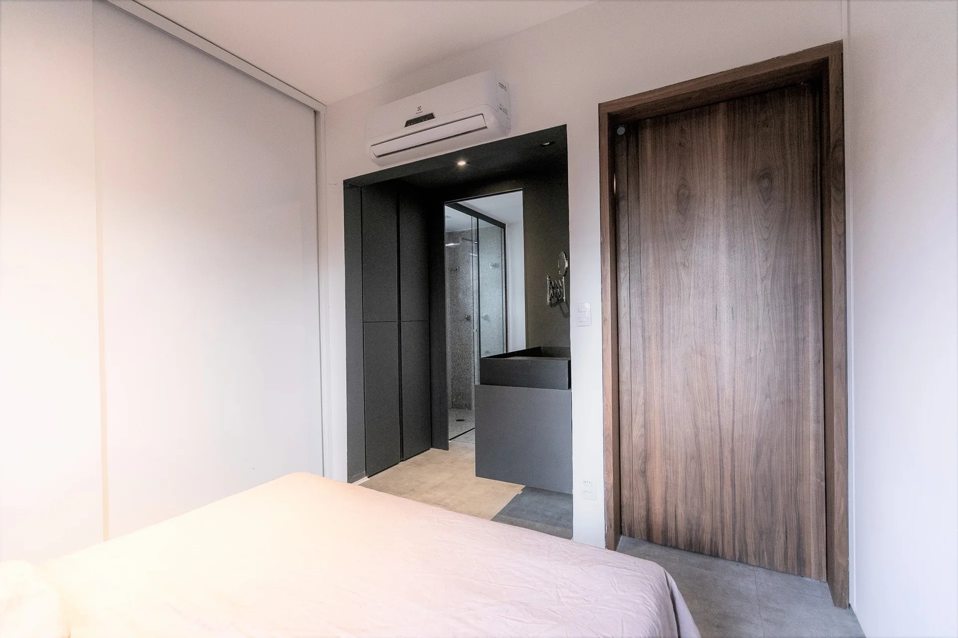 A modern bedroom with light bedding features a tall wood grain door and an open entryway to a dark grey adjoining bathroom.