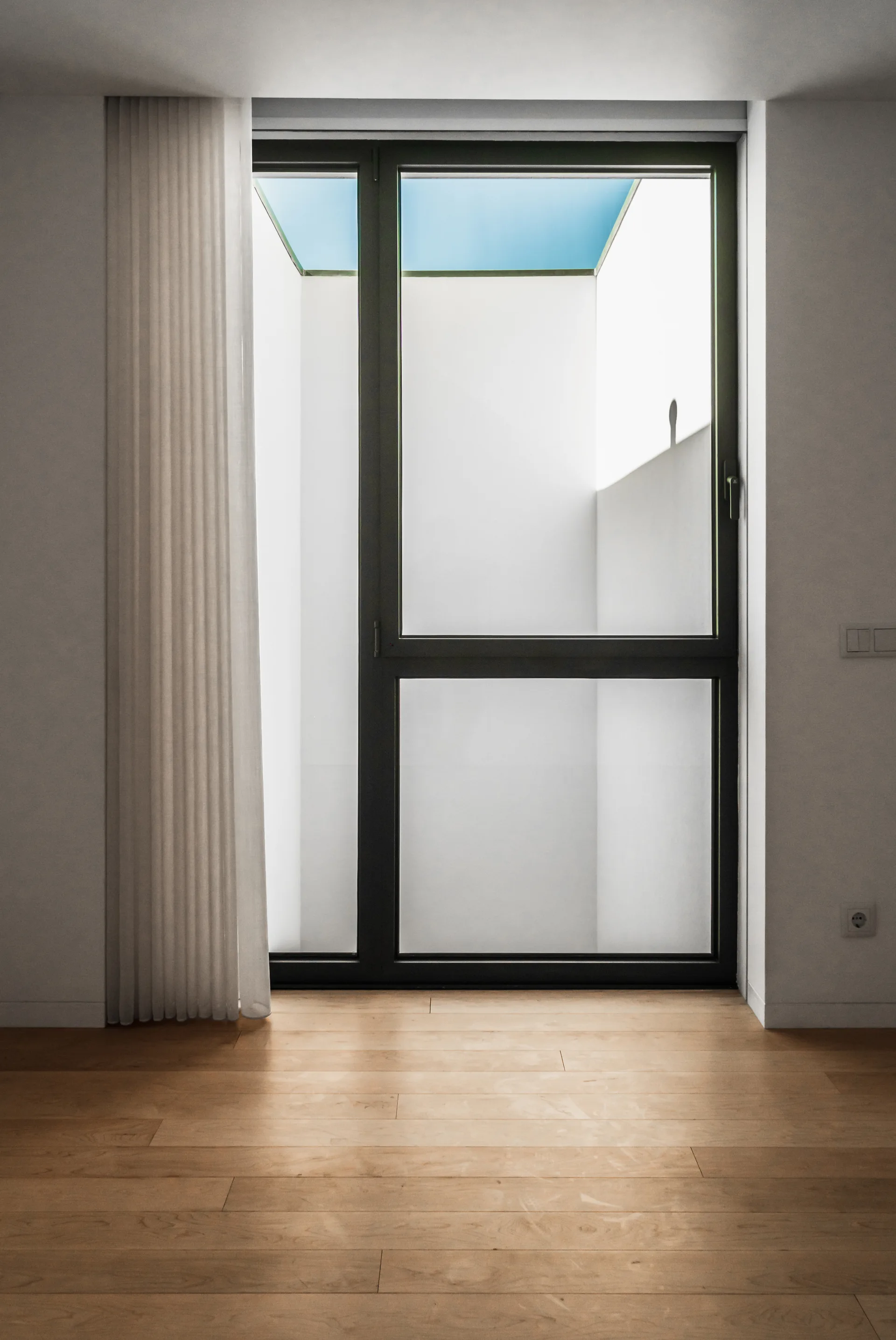 A minimalist room features light hardwood floors, white walls, and a dark-framed glass door opening to a bright lightwell with a sheer white curtain.