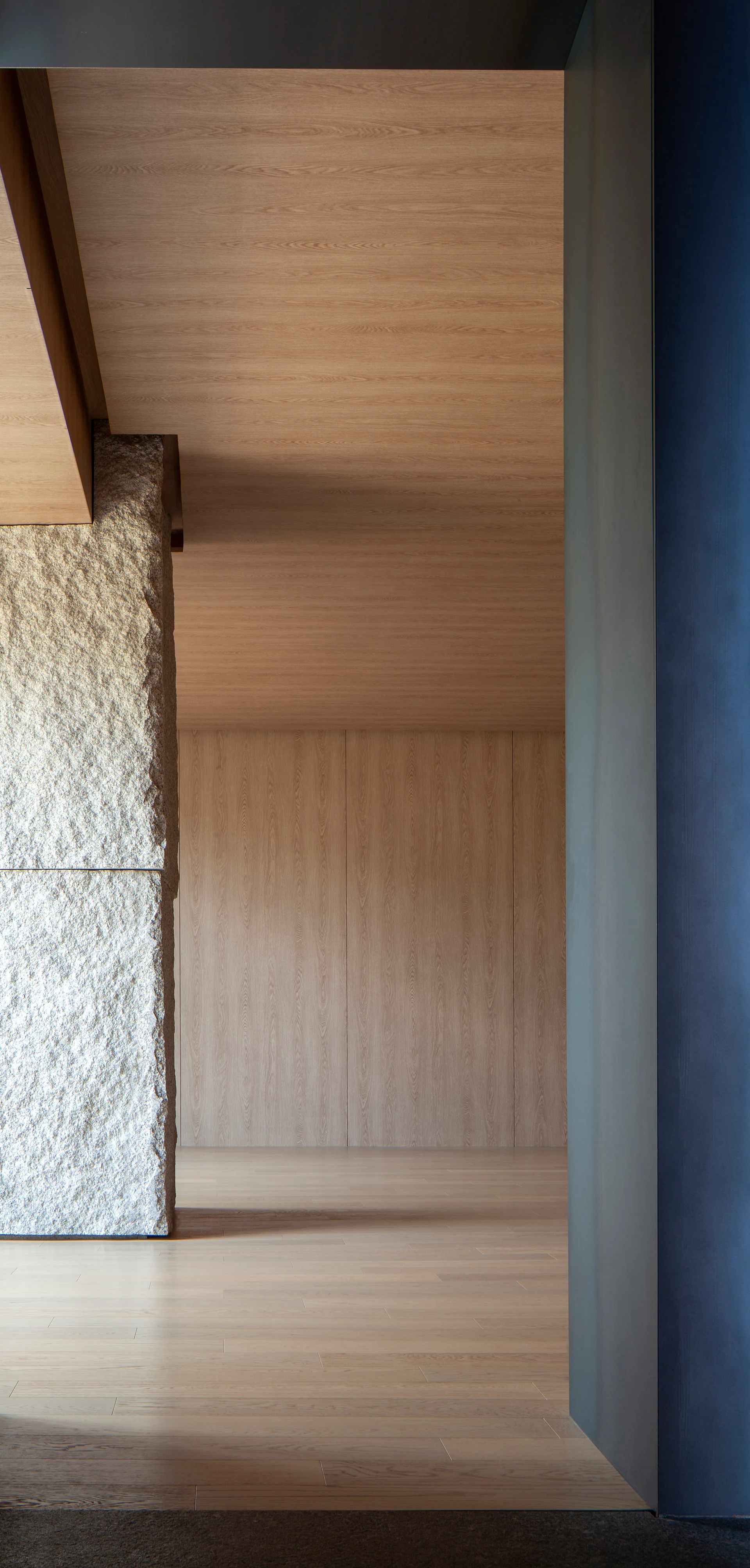 A minimalist transitional hallway displays a rough stone pillar contrasted by smooth light wood paneling on the walls, ceiling, and floor.