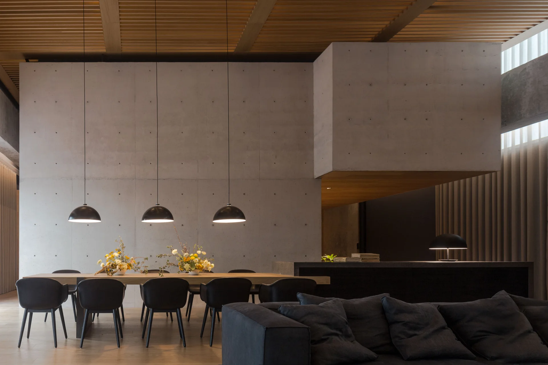 A modern dining area features black chairs around a wooden table beneath three dome pendant lights, backed by a large grey concrete wall.