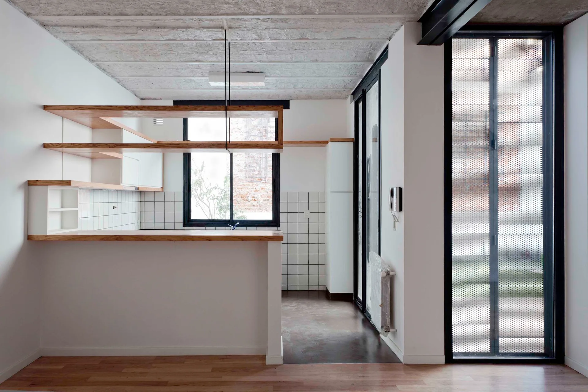 A minimalist kitchen features a white tile backsplash, light wood open shelves, an island counter, and an exposed concrete ceiling.