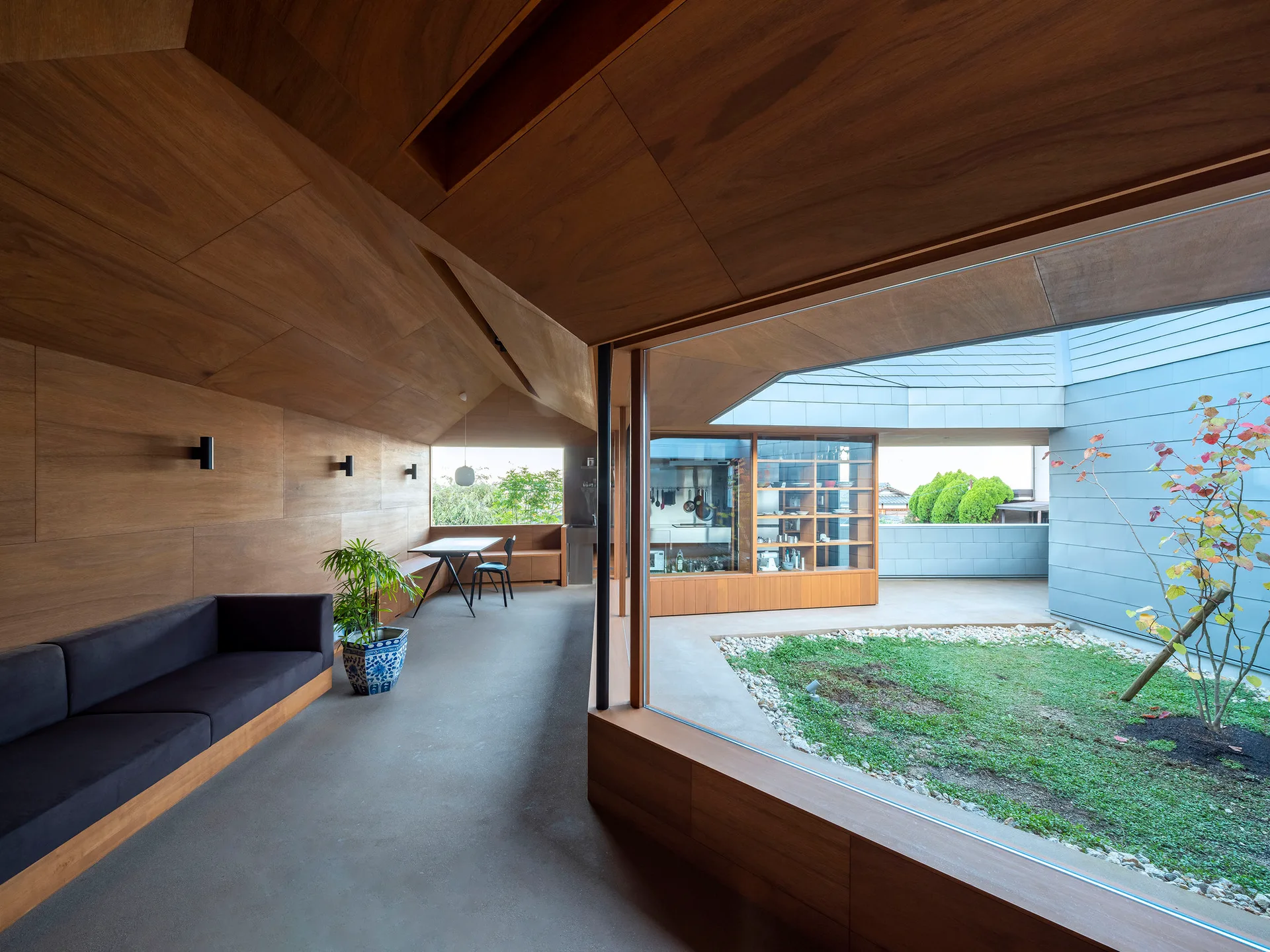 A modern living area featuring warm wood-paneled walls and ceiling, a dark built-in sofa, and large windows looking out onto a grassy courtyard.