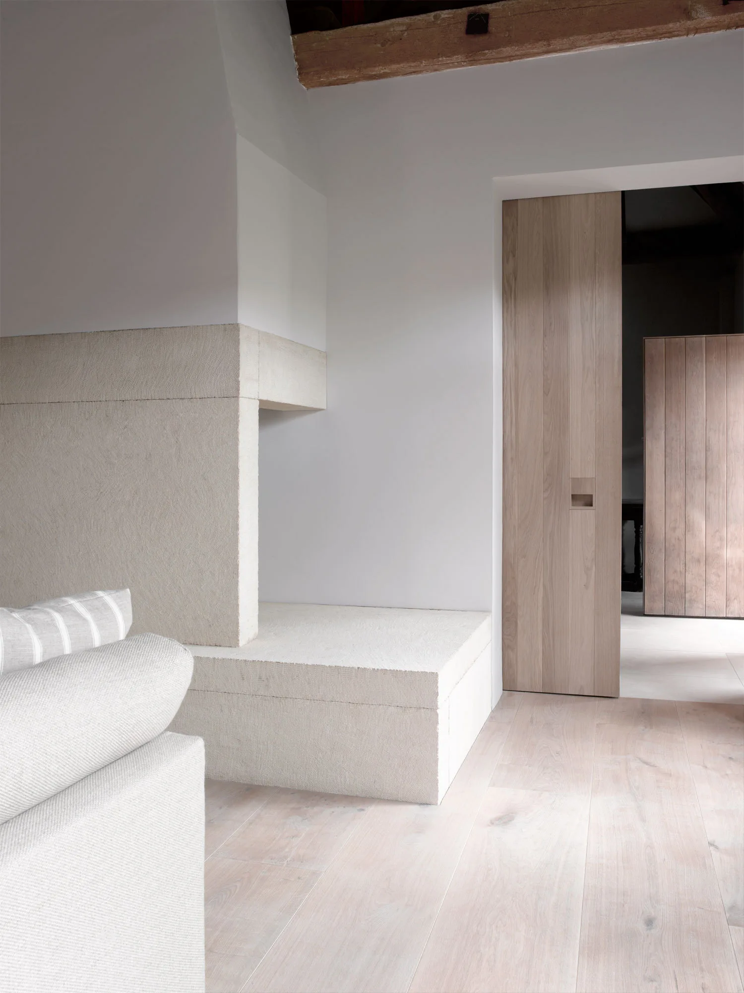 A minimalist room features a textured beige stone fireplace hearth next to a tall, light wood sliding pocket door and matching wood flooring.