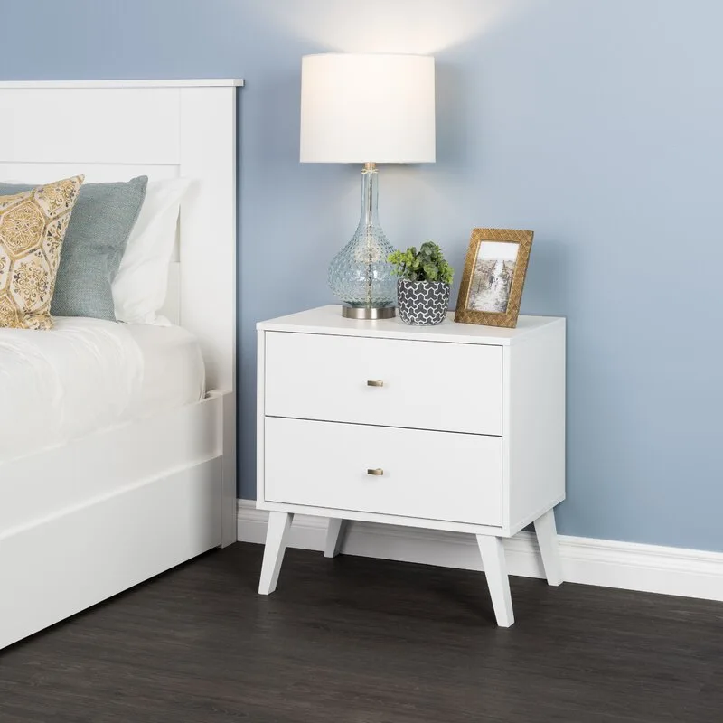 A white two-drawer nightstand with tapered legs stands next to a bed against a blue wall, holding a blue glass lamp and a gold frame.