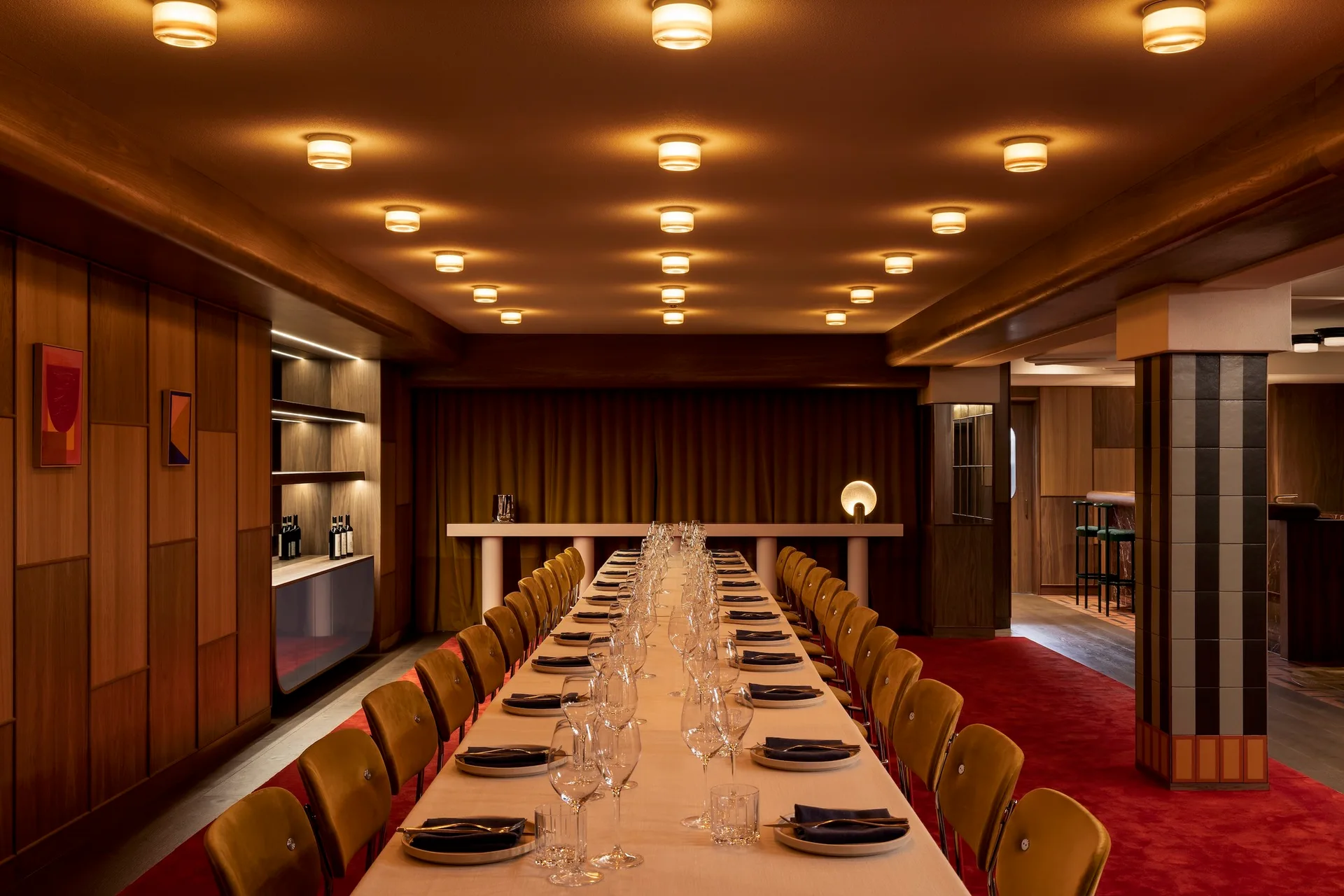 A long dining table set with glasses and plates stands on a vibrant red carpet beneath a grid of glowing flush-mount ceiling lights.