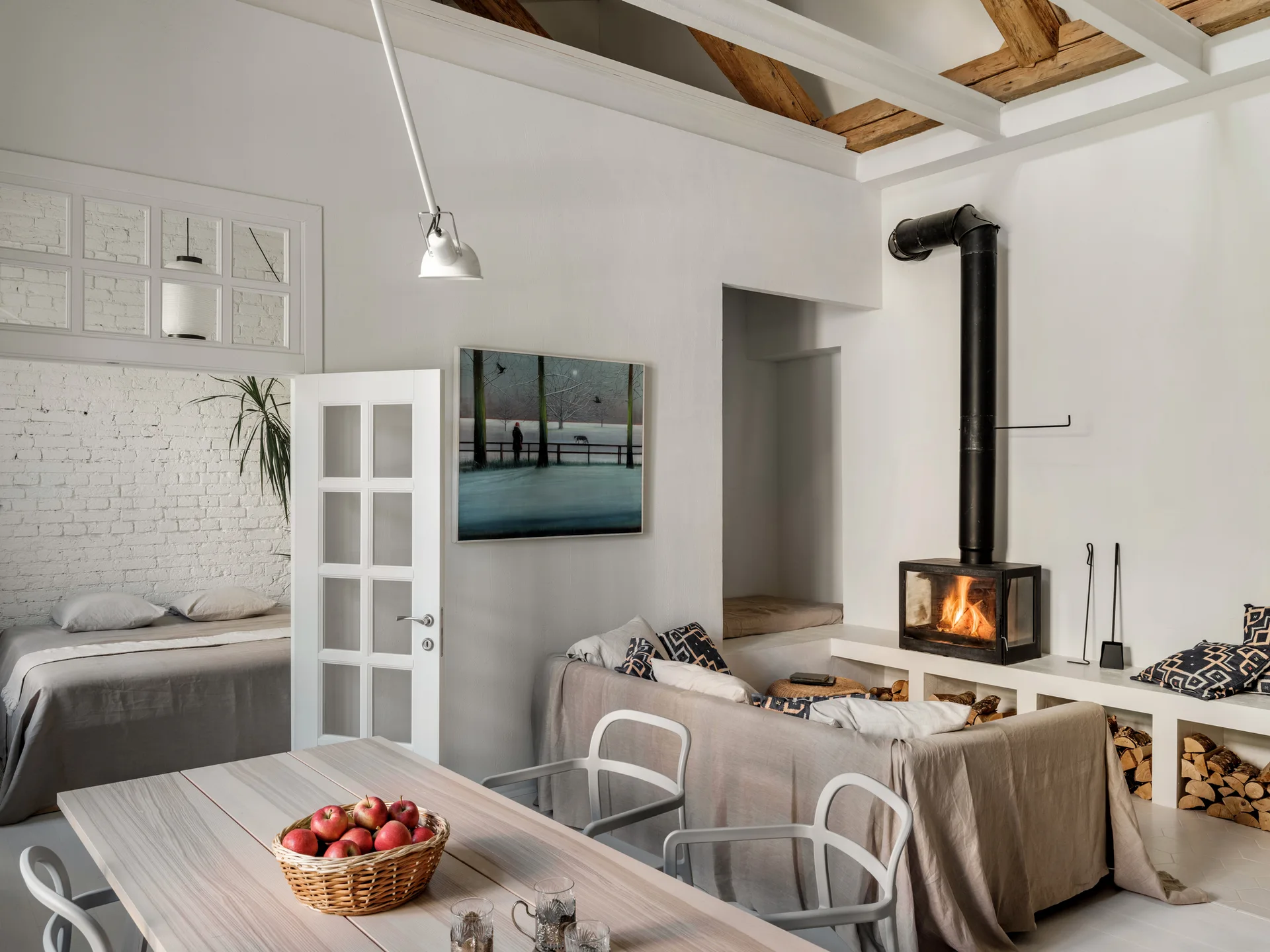 A light-filled open living space features a modern black fireplace, a light wood dining table, and exposed ceiling beams.