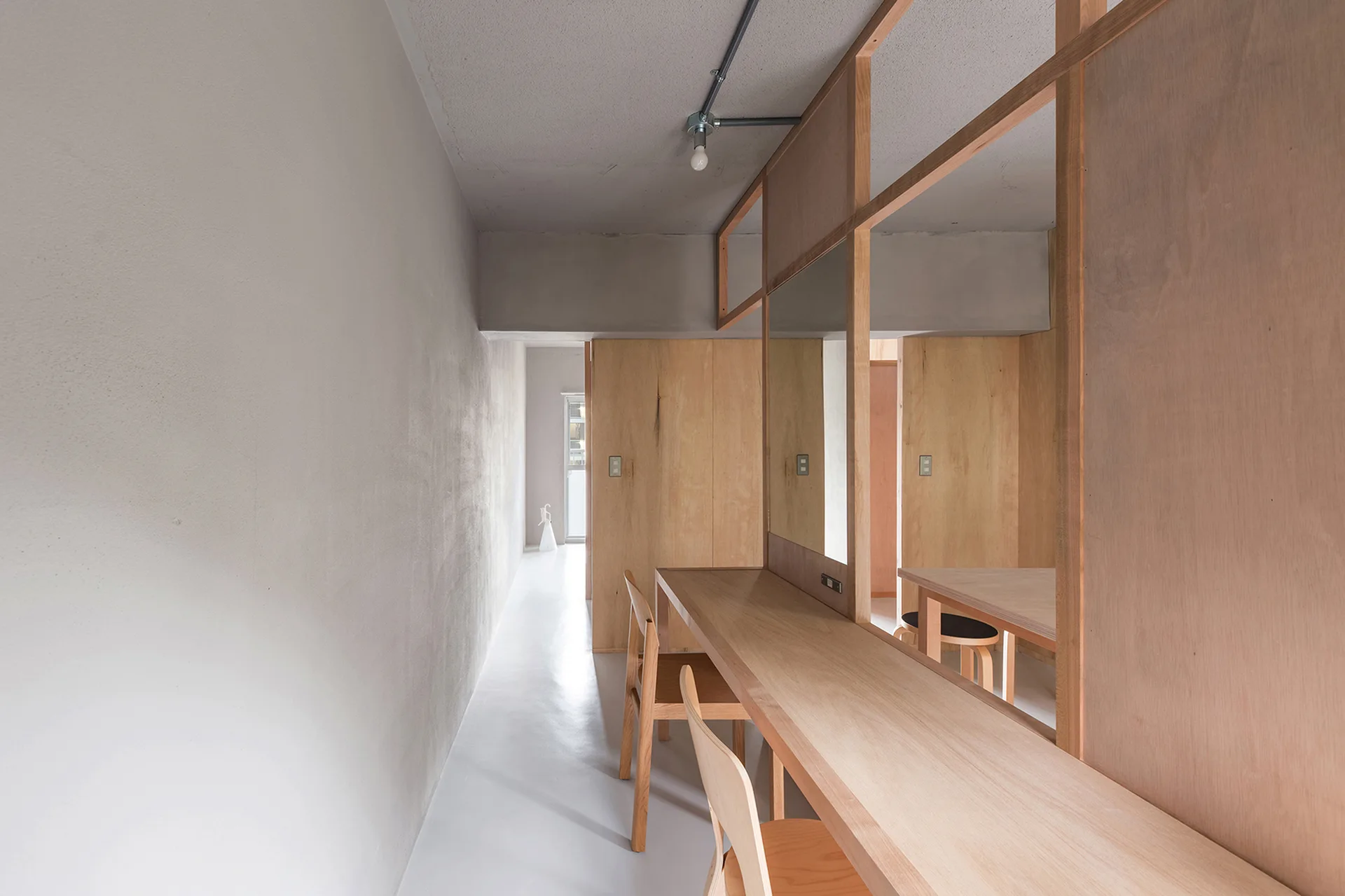 A long built-in wooden desk with matching chairs is integrated into a plywood room partition with a mirror inside a minimalist grey corridor.
