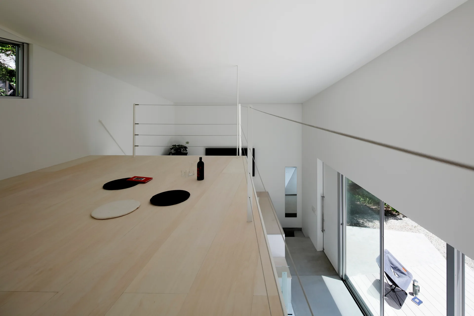 A minimalist mezzanine with light wood flooring and a thin white wire railing overlooks a lower level with a large sliding glass door.