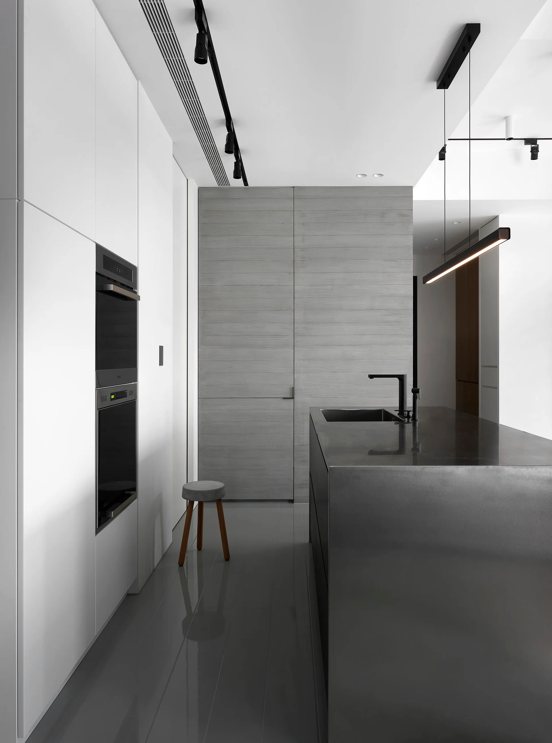 A minimalist kitchen features a dark metallic island with a black faucet, white handleless cabinetry with integrated ovens, and a gray wood accent wall.