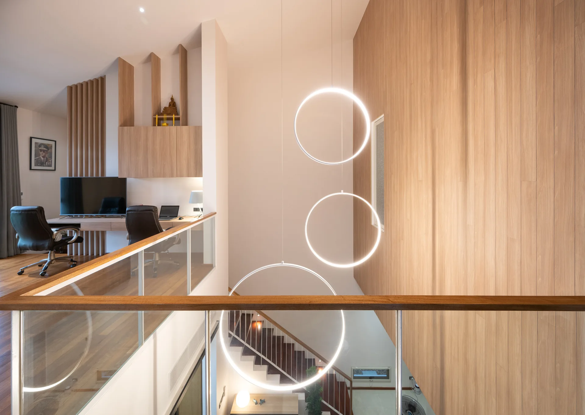 A lofted home office with a wood-planked accent wall overlooks a staircase, featuring glass railings and three suspended circular ring pendant lights.