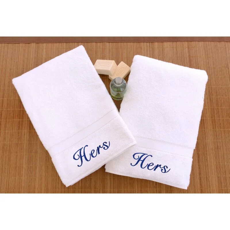 Two white cotton bath towels with blue embroidered text reading Hers are displayed on a woven brown mat alongside two bars of soap.