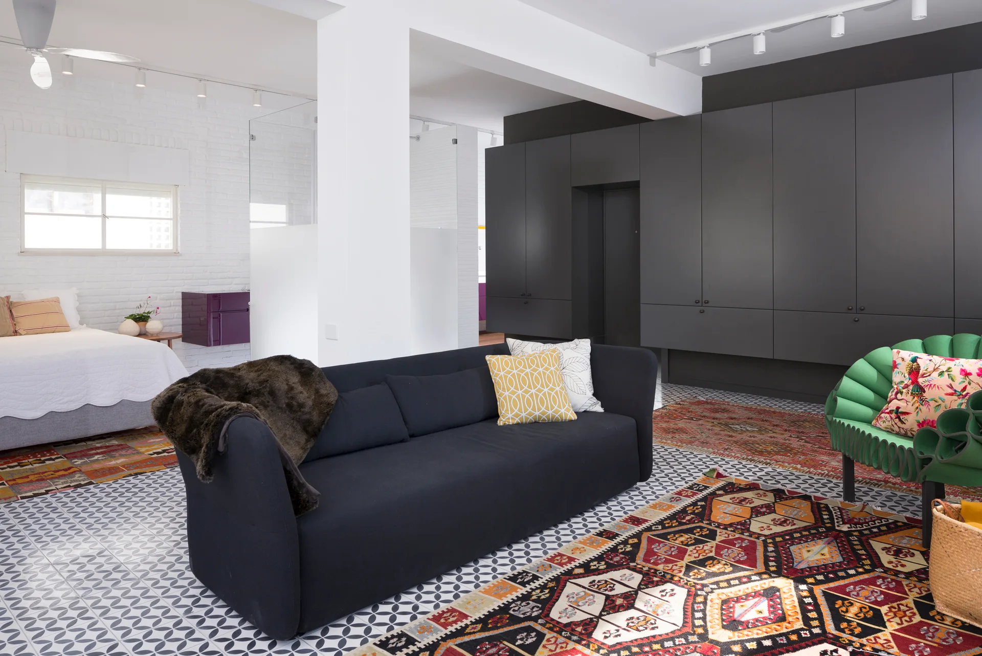 An open-plan living room and bedroom featuring a dark sofa, patterned rugs, graphic floor tiles, and a wall of charcoal built-in cabinets.