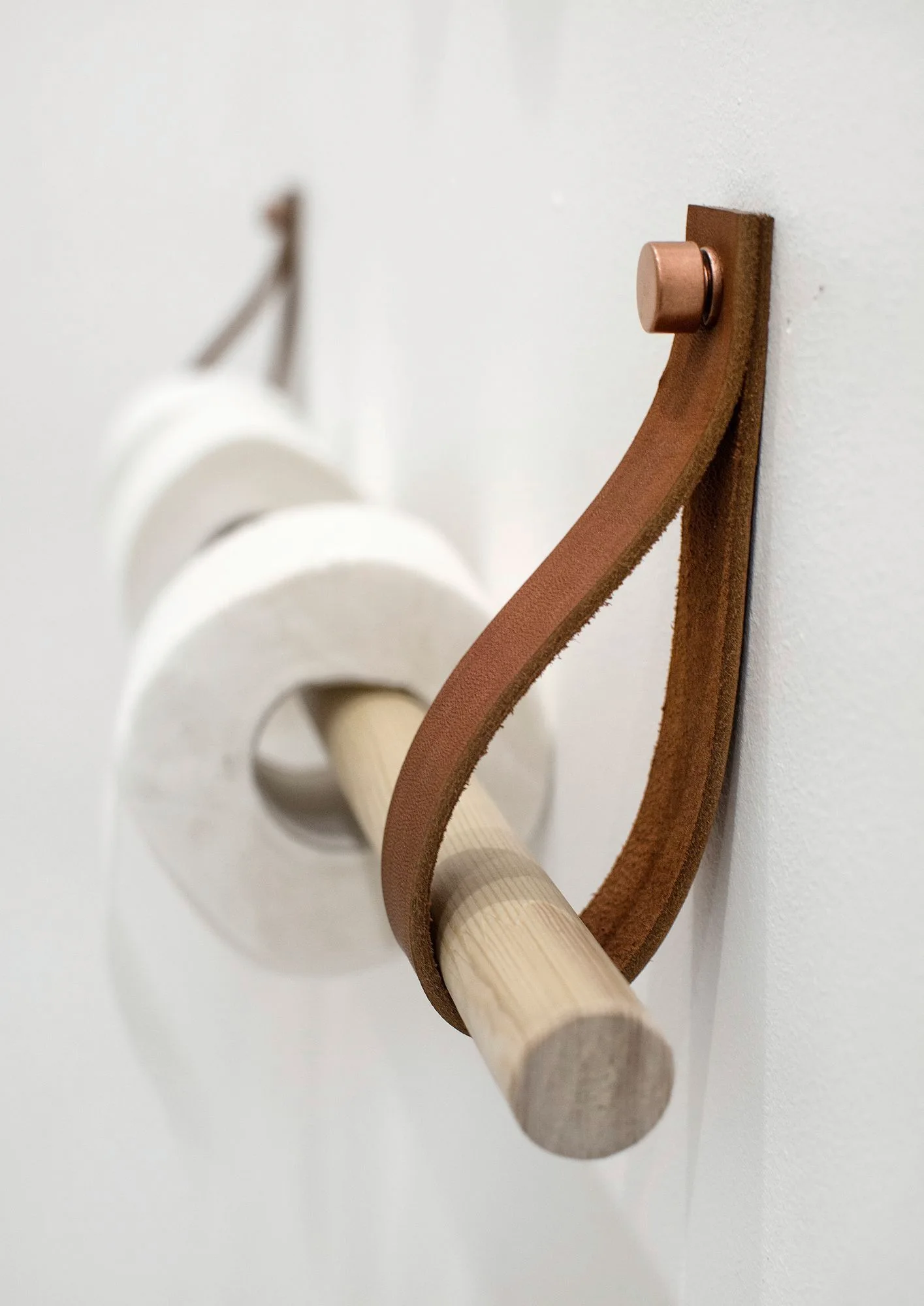 A minimalist toilet paper holder made of a light wood dowel and brown leather straps mounted on a white wall.