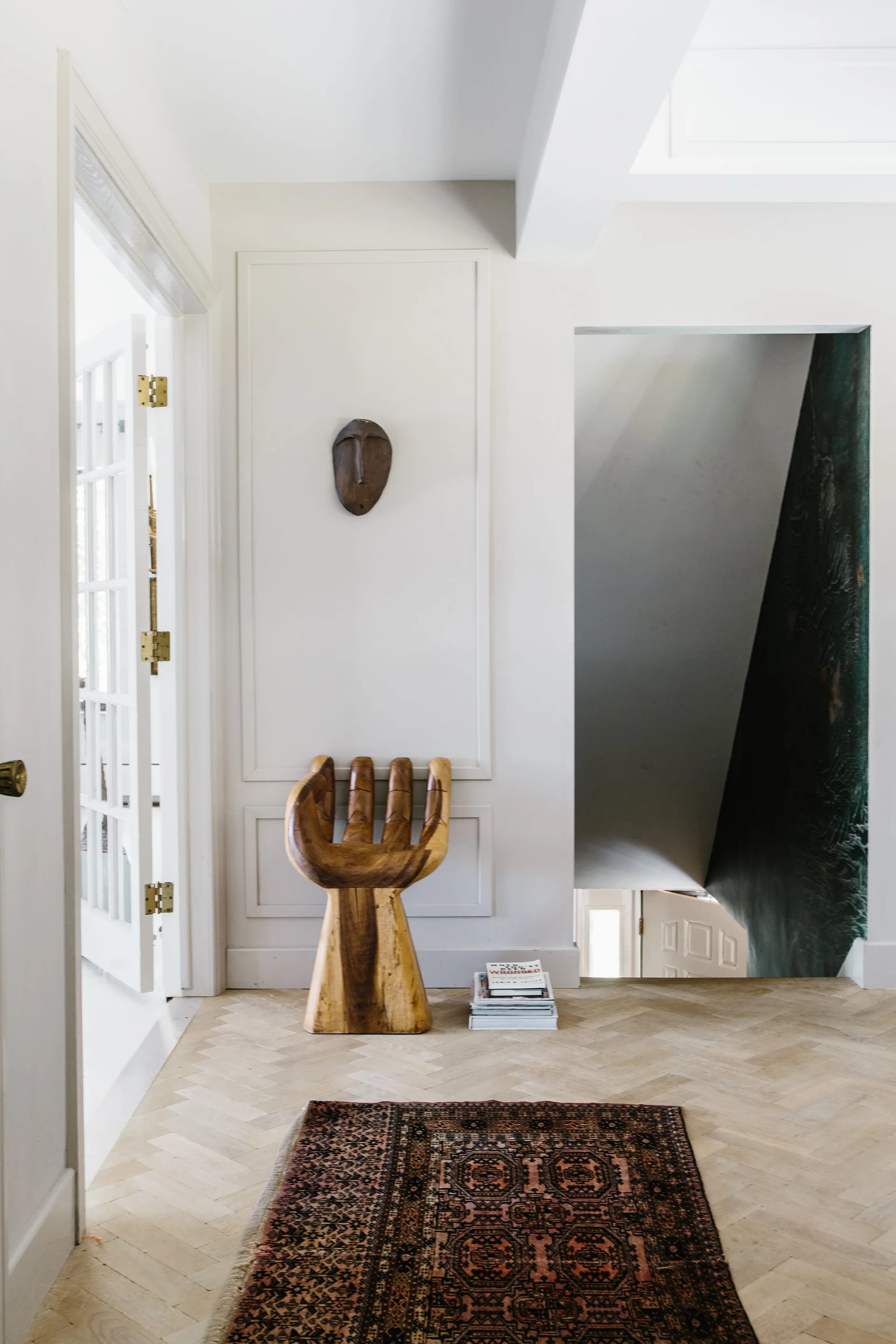 A bright hallway features herringbone wood flooring, a sculptural hand-shaped wooden chair, a wall-mounted mask, and a small patterned rug.