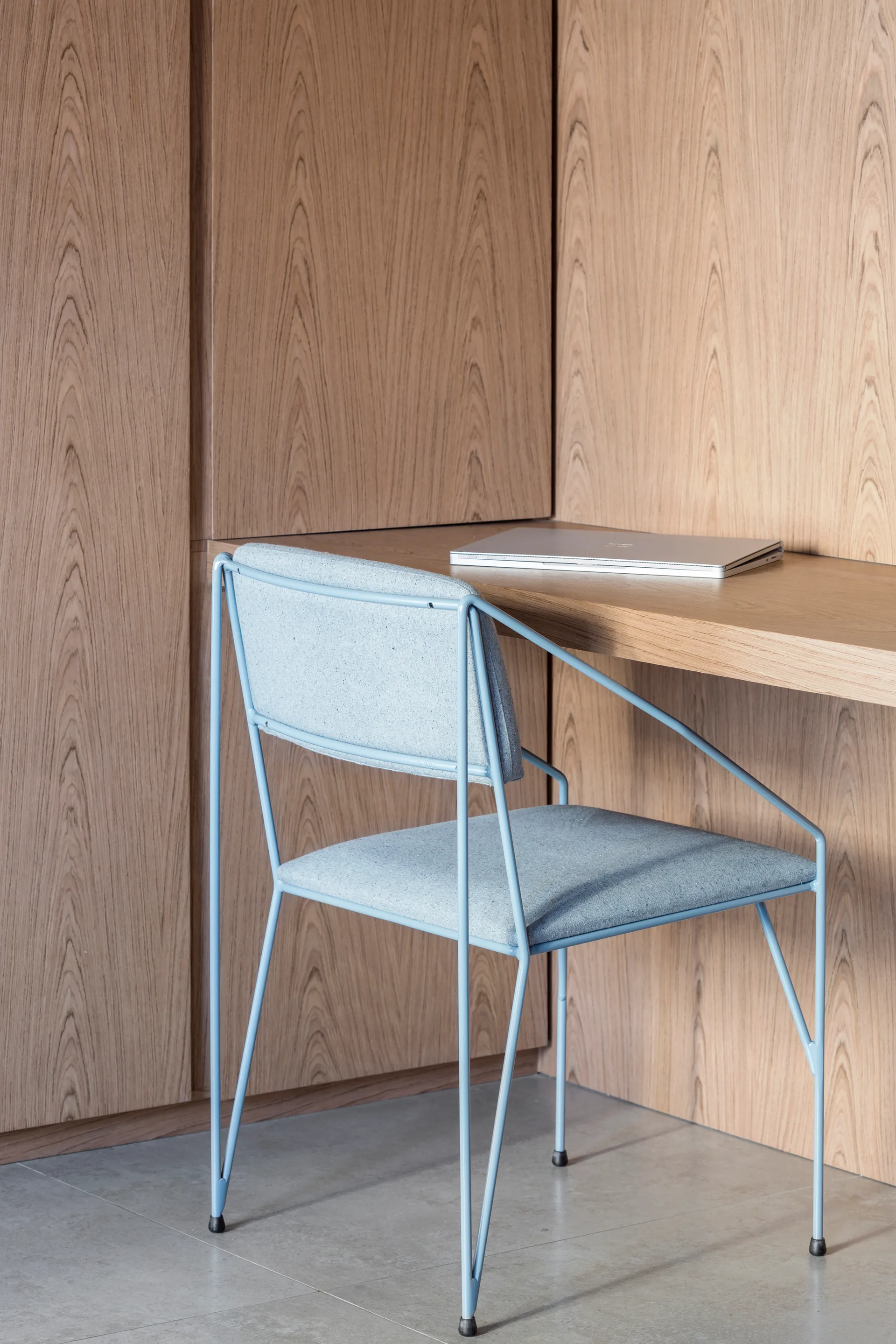 A minimalist home office nook features light wood paneling on the walls and built-in desk, paired with a light blue metal-frame chair.