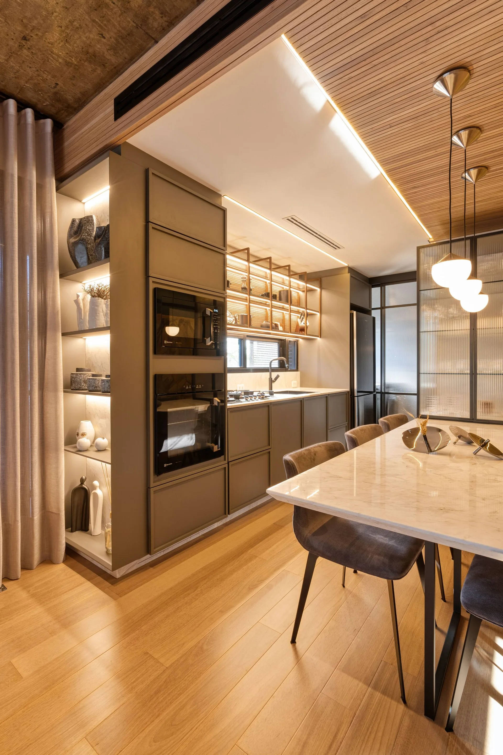 A modern kitchen features matte olive green cabinetry, a marble dining table with grey chairs, and a light wood slatted ceiling detail.