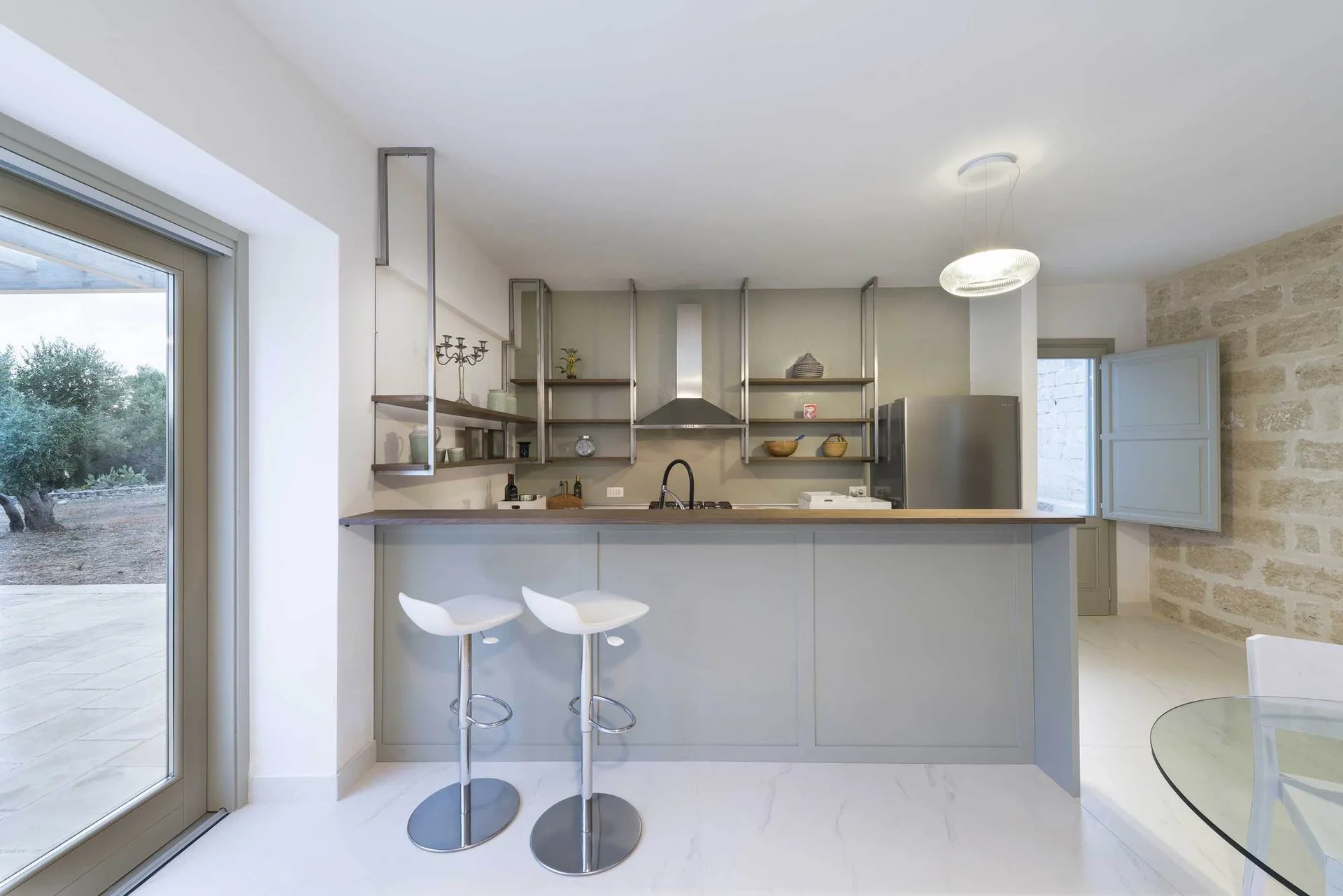 A modern kitchen features a light gray island with white bar stools, open metal shelving on the wall, and an exposed stone block accent wall.
