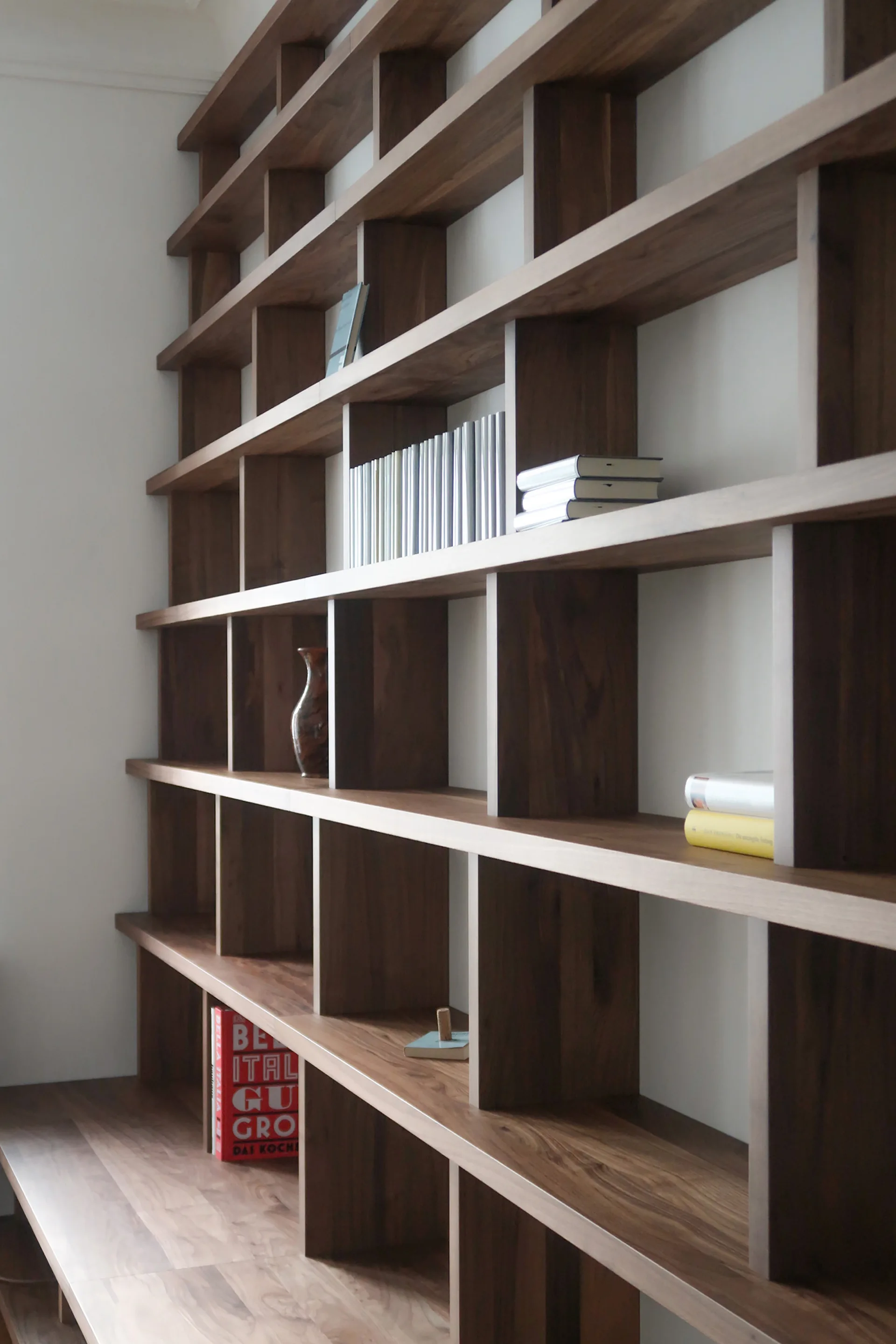 A dark wood open grid bookshelf is mounted against a white wall with minimal books and decorative accessories.