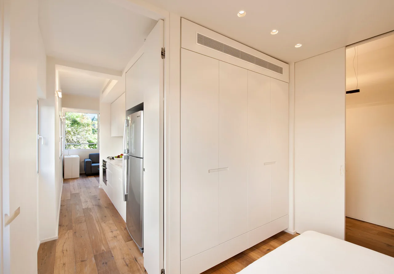 White seamless built-in storage cabinets stand opposite a narrow galley kitchen with a stainless steel refrigerator and wood flooring.