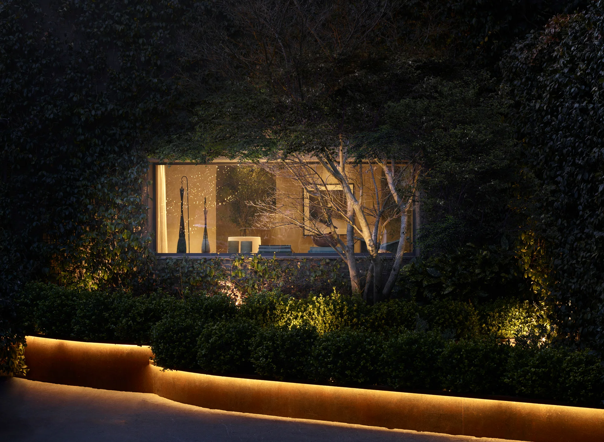 A dark night garden features a rectangular window showing a warm interior, surrounded by dense green ivy and a glowing LED strip along a planter.