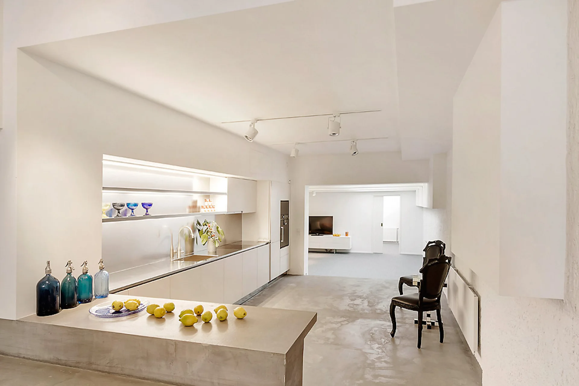 A minimalist white kitchen features concrete-like floors, a long island, under-cabinet lighting, and two black classic chairs.