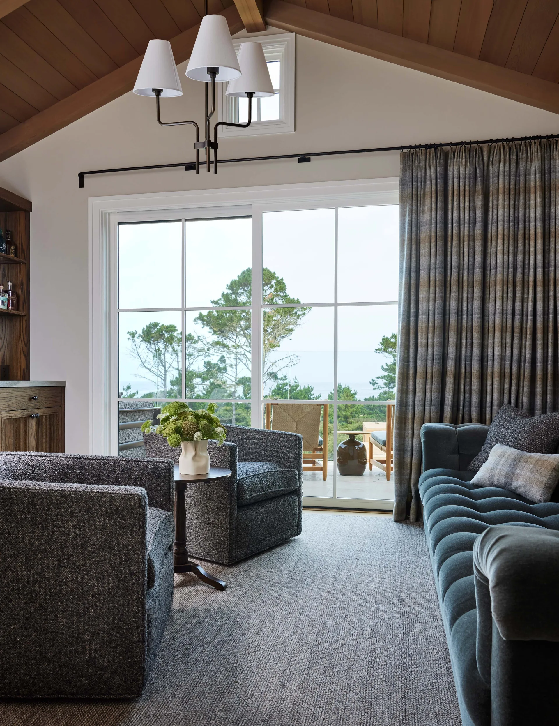 A living room features a blue tufted sofa, two gray tweed armchairs, and a large sliding glass door opening to a deck under a vaulted wood ceiling.