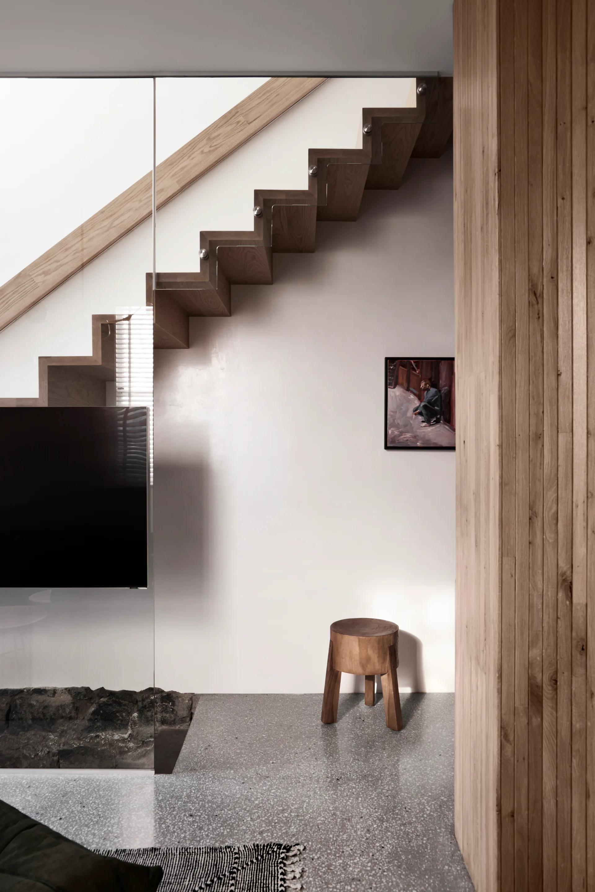 A floating wooden staircase with a glass railing is shown next to a terrazzo floor, a small wood stool, and a vertical wood-paneled wall.