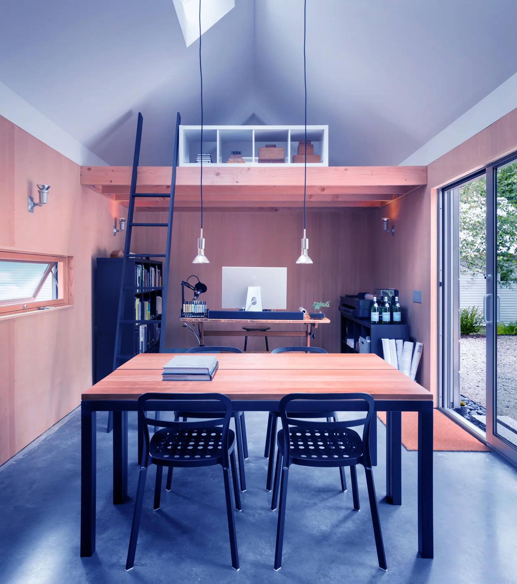 A minimalist wood-paneled home studio with a vaulted ceiling, concrete floor, central wooden table, and a loft storage space with a black ladder.