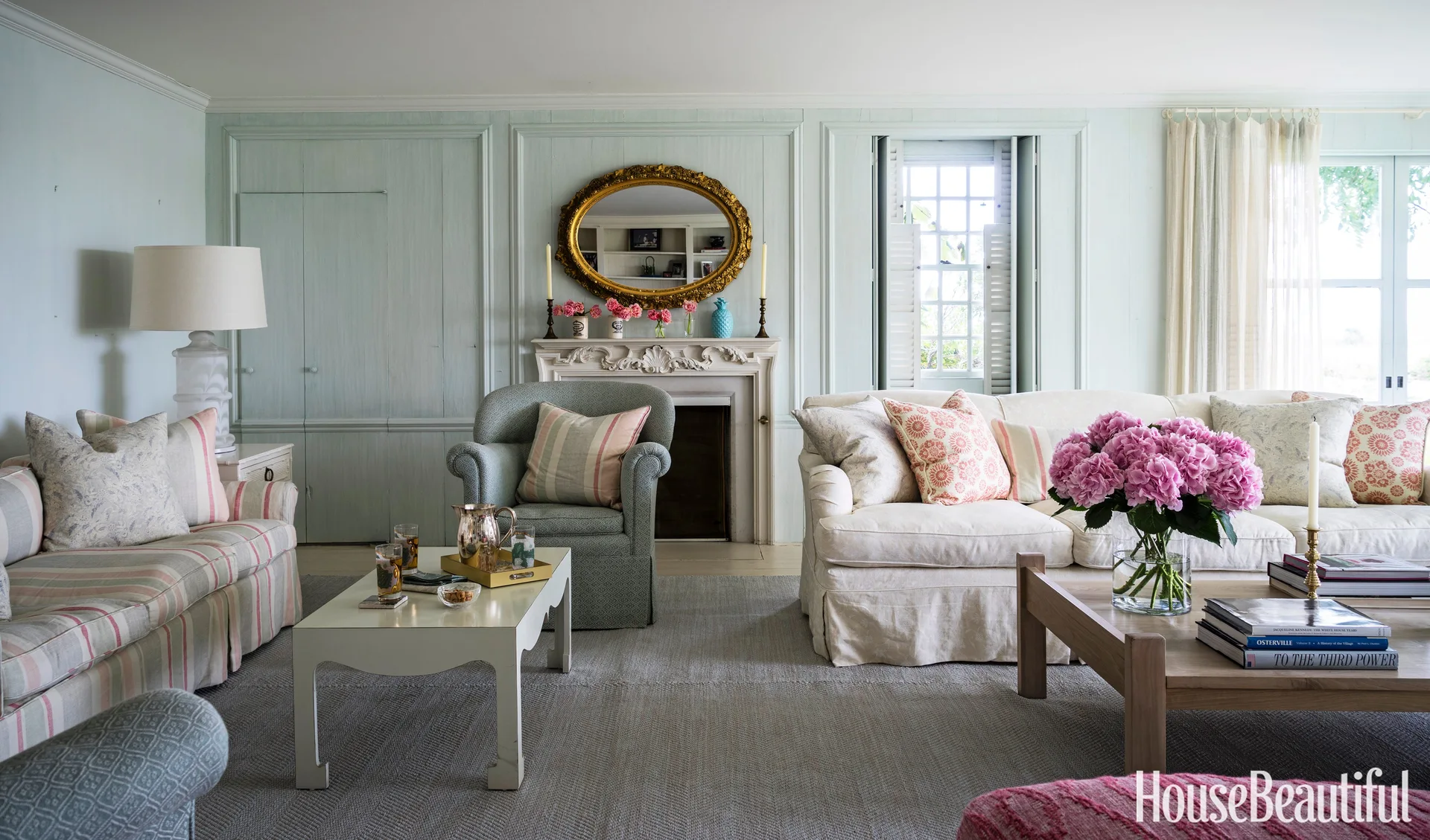A living room featuring light blue paneled walls, a fireplace with an ornate gold mirror, two coffee tables, and comfortable slipcovered sofas.