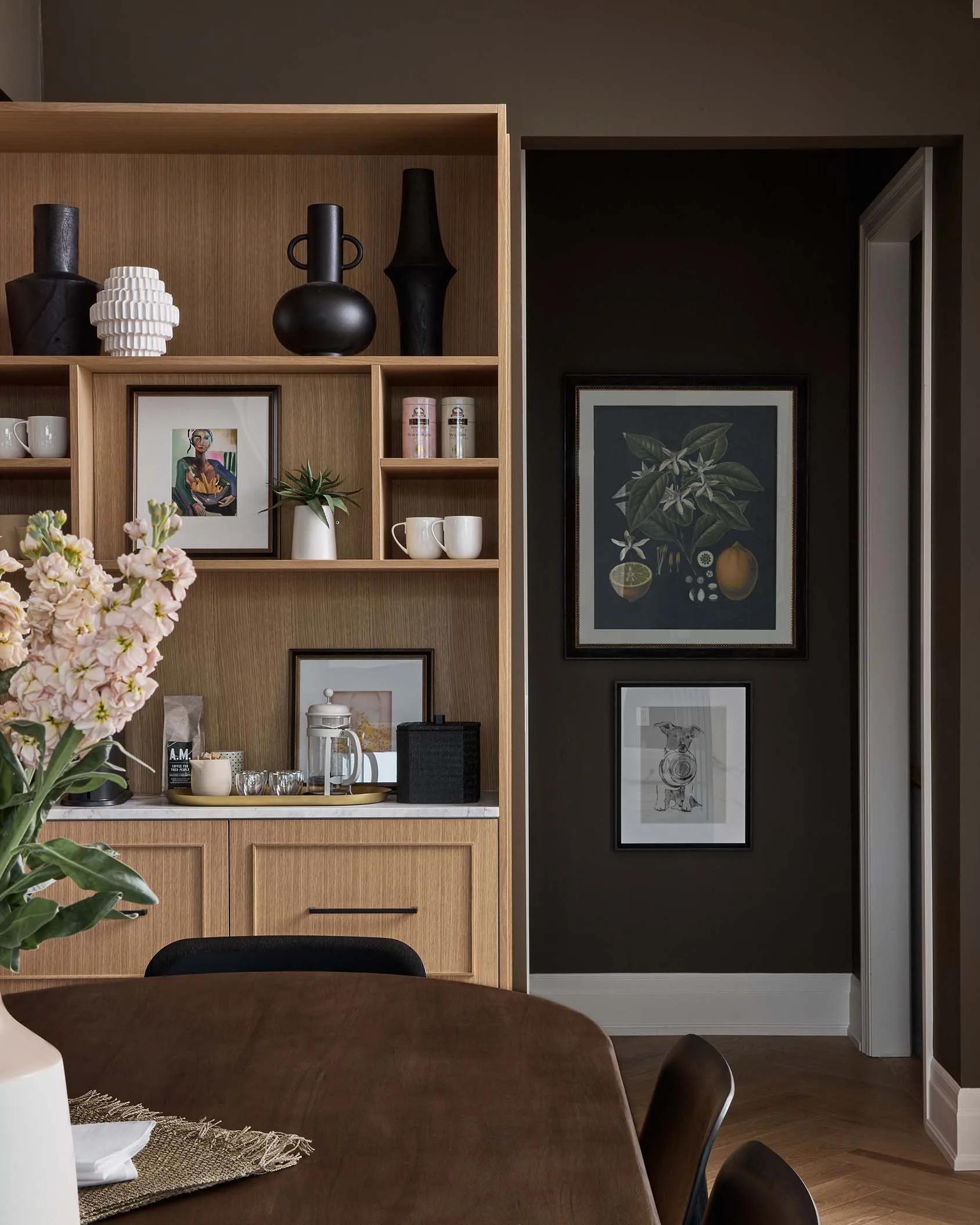 A light oak built-in cabinet with open shelving sits next to dark brown walls decorated with framed prints in a dining room.