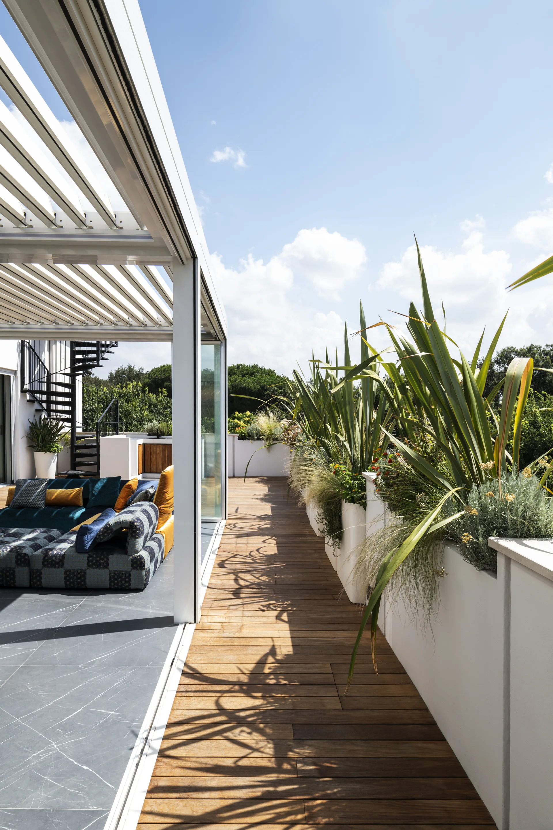 An outdoor rooftop terrace with a modern modular sofa under a louvered pergola next to a row of tall white planters.