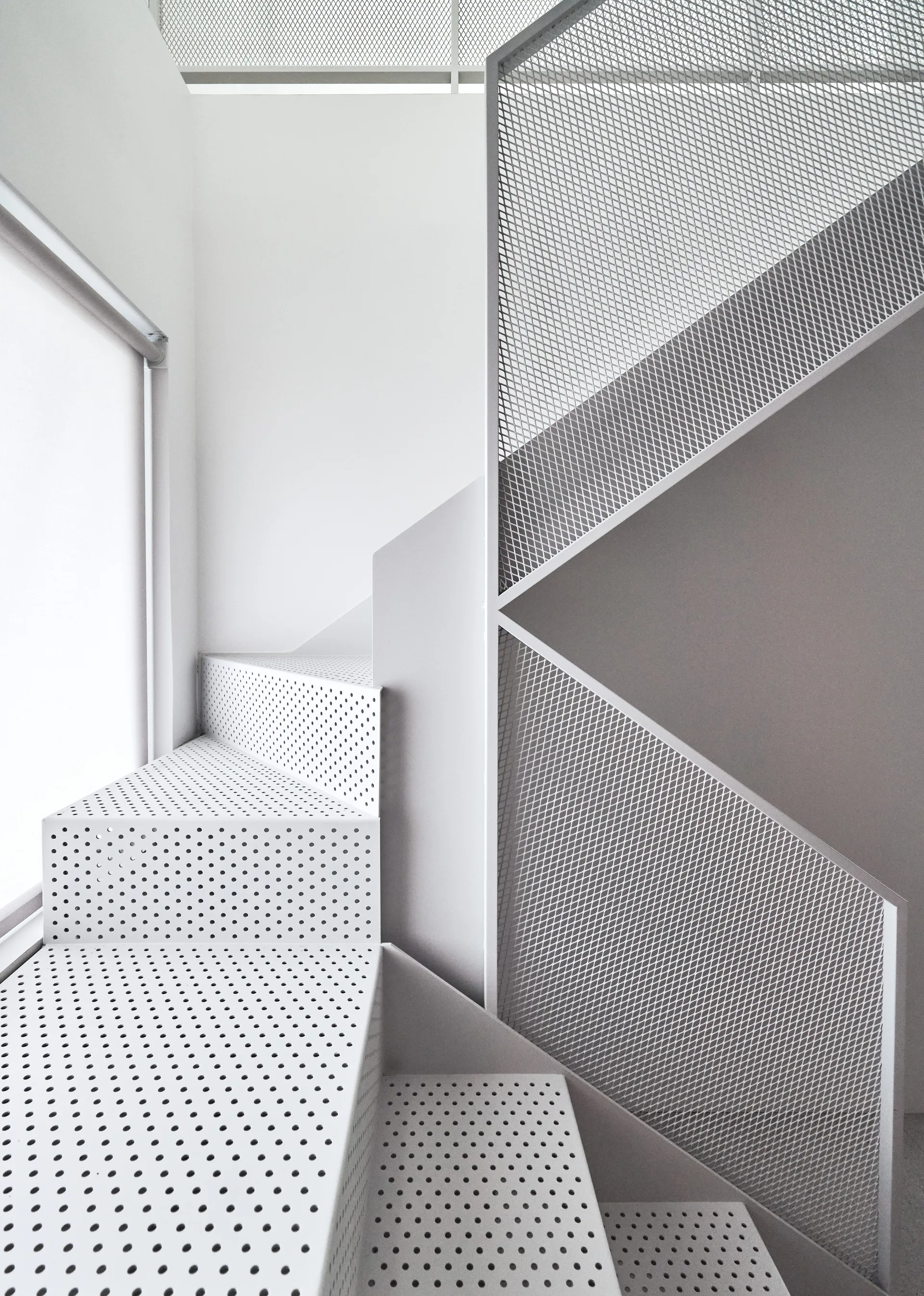 A modern white staircase featuring perforated metal treads and a geometric metal mesh railing next to a bright window.