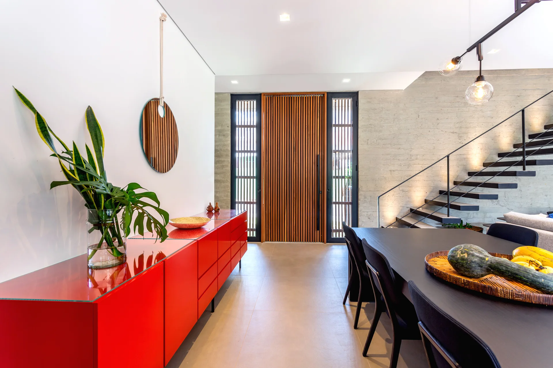 A modern entryway features a long glossy red console table, a round hanging mirror, a vertical wood slat door, and a floating staircase against a concrete wall.